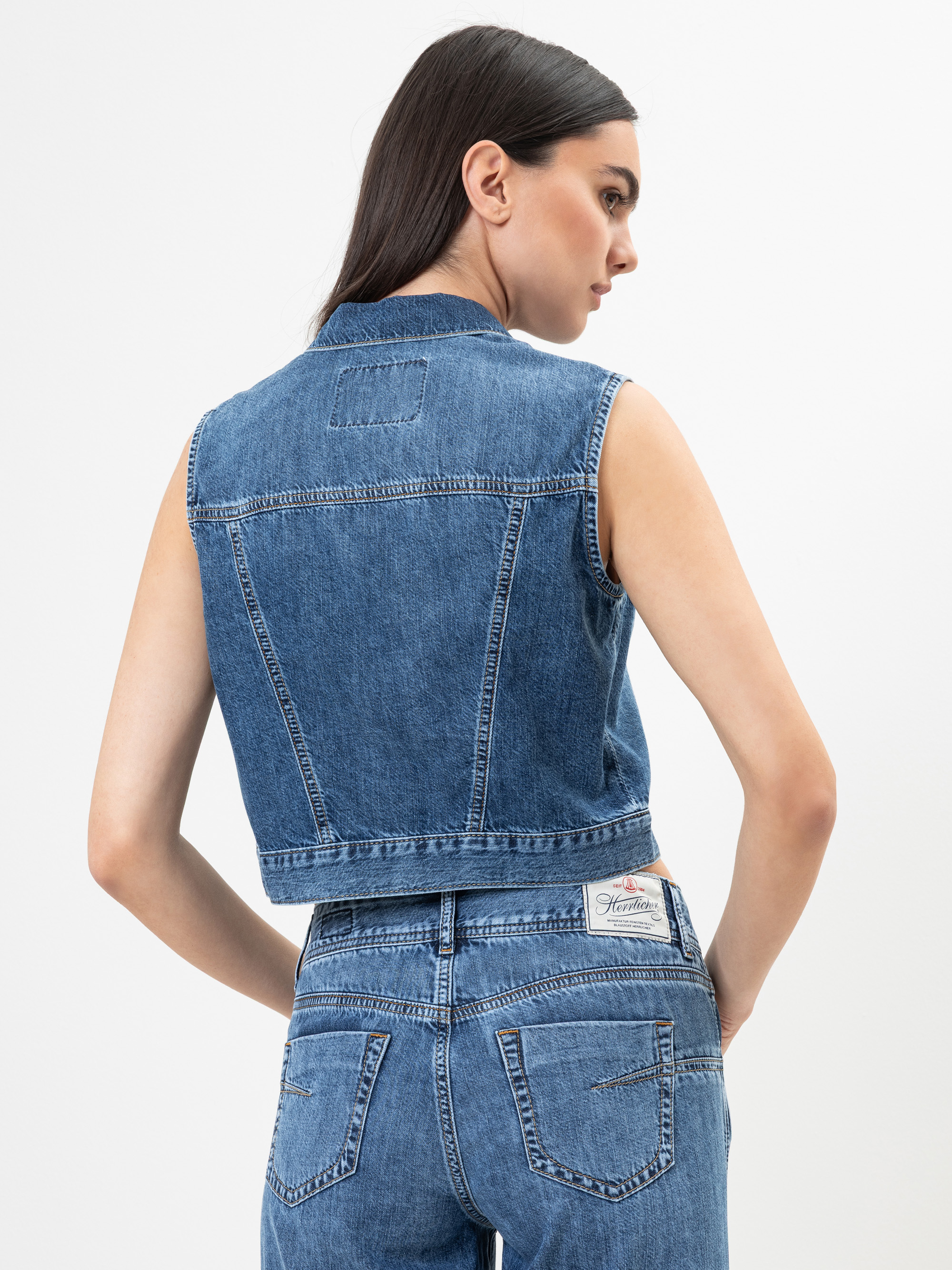 Eine Frau steht mit dem Gesicht zur Wand und trägt eine ärmellose blaue Jeansweste und dazu passende Jeans mit hoher Taille. Der Jeansstoff hat eine klassische mittlere Waschung, und sie steht vor einem schlichten weißen Hintergrund.