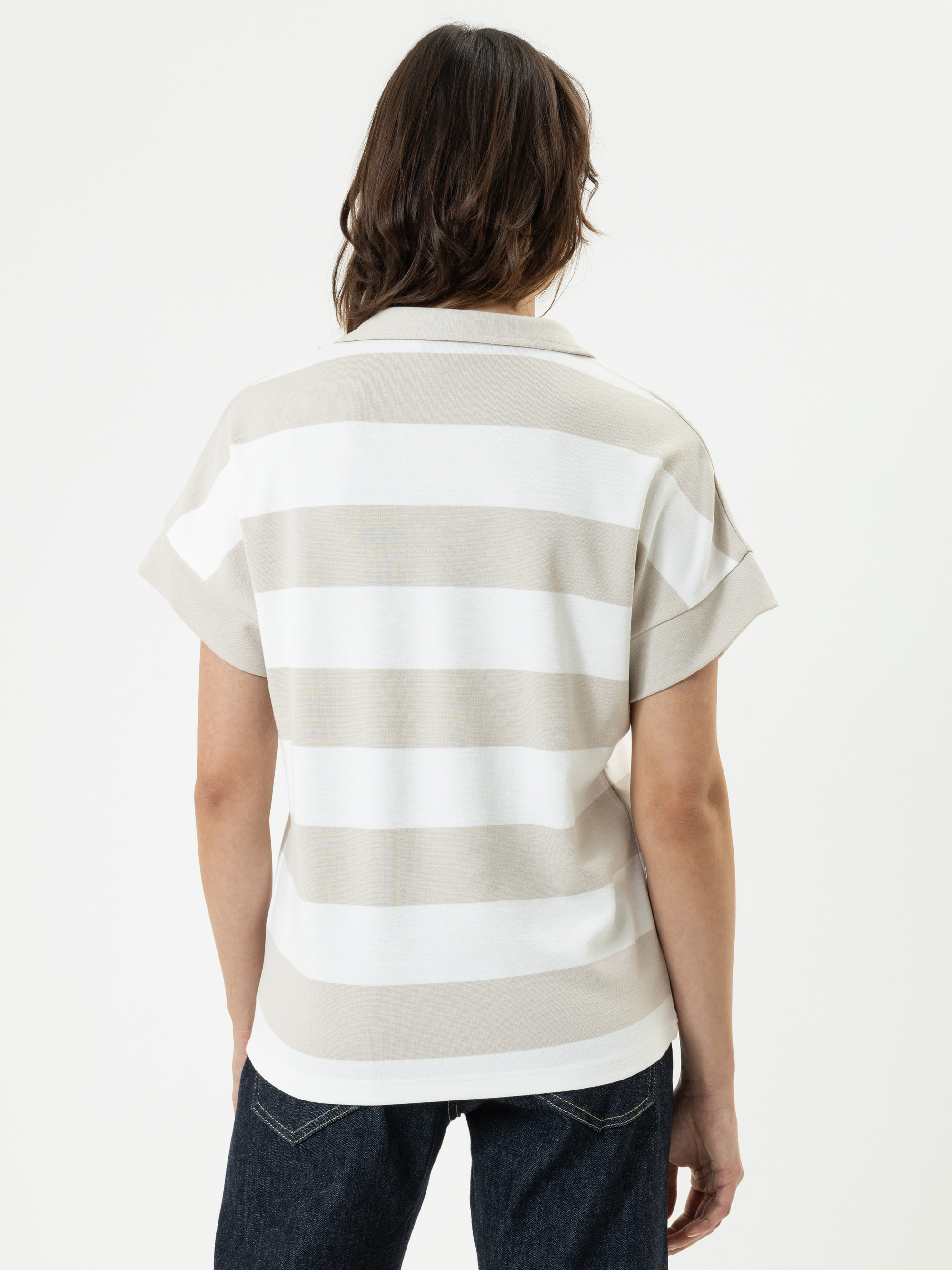 Eine Person mit schulterlangen braunen Haaren ist von hinten zu sehen. Sie trägt ein kurzärmeliges, locker sitzendes Hemd mit breiten beigen und weißen Querstreifen und eine dunkle Jeans vor einem schlichten weißen Hintergrund.