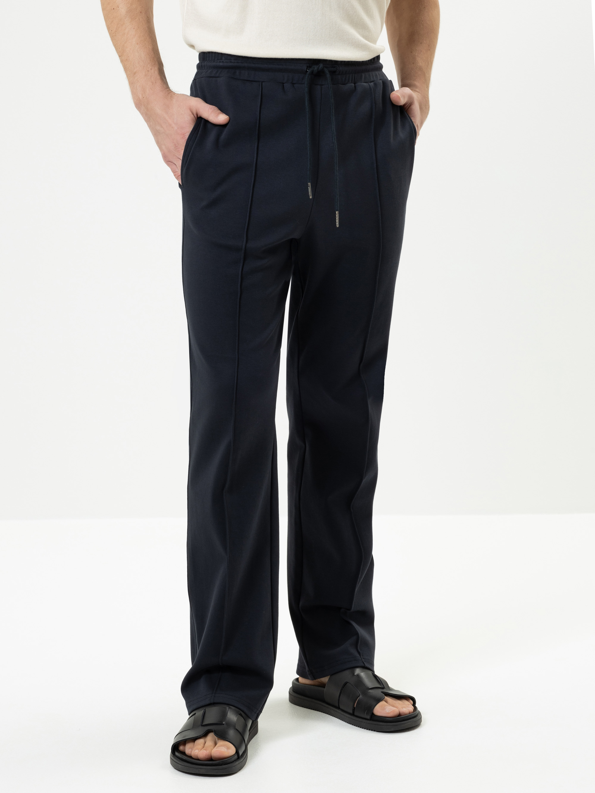 Eine Person, die eine marineblaue Hose und schwarze Sandalen trägt, steht mit den Händen in den Taschen vor einem schlichten weißen Hintergrund.