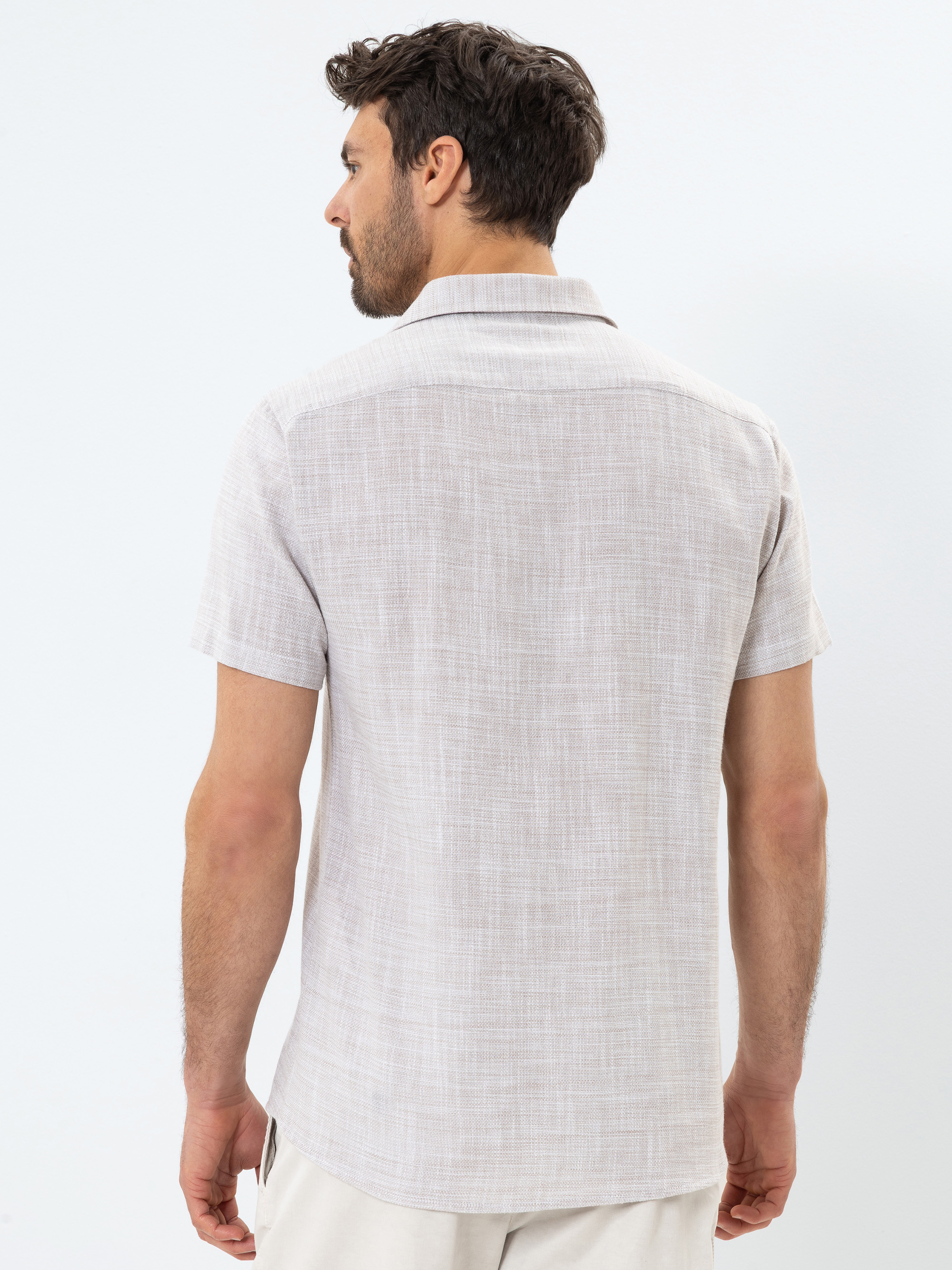Ein Mann mit kurzen dunklen Haaren und einem Bart wird von hinten gezeigt. Er trägt ein hellgraues, kurzärmeliges, strukturiertes Button-up-Hemd und eine helle Hose. Der Hintergrund ist einfarbig weiß.