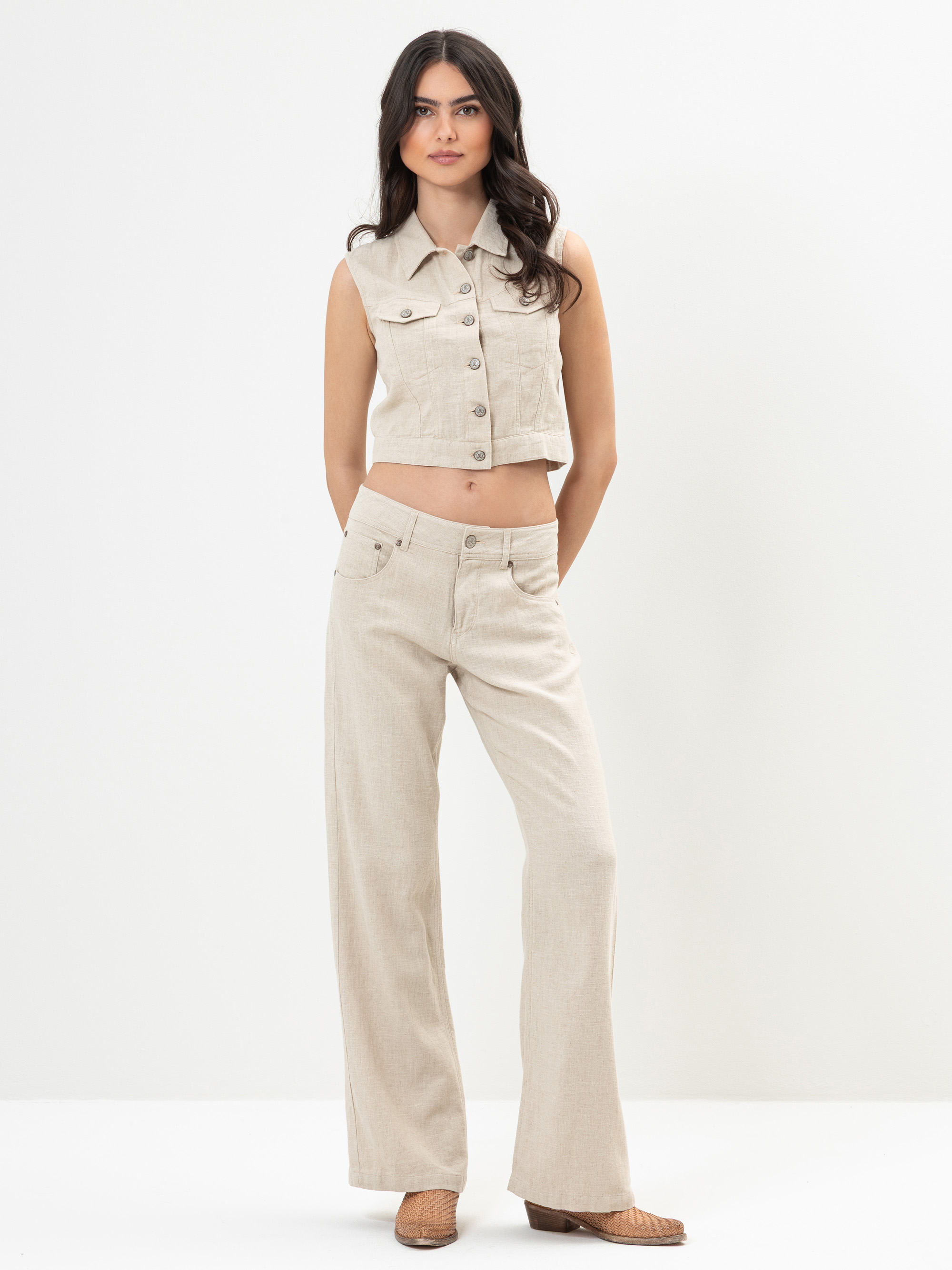 Eine Frau steht vor einem schlichten weißen Hintergrund und trägt ein ärmelloses, geknöpftes beigefarbenes Oberteil und eine passende Hose mit hoher Taille und weitem Bein sowie hellbraune Schuhe. Sie hat langes, dunkles Haar und einen neutralen Gesic