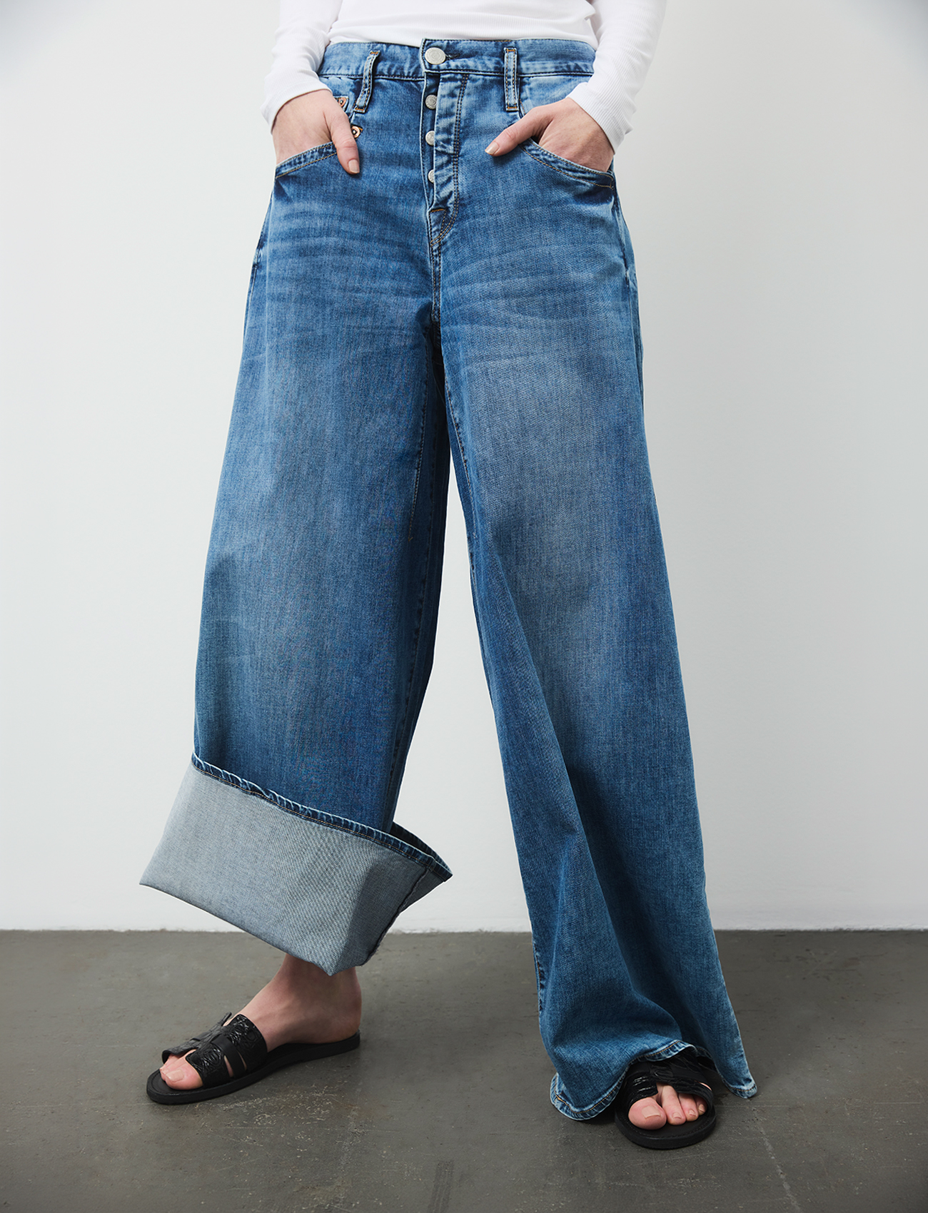 Eine Person steht vor einer schlichten Wand und trägt eine blaue Jeans mit weitem Bein, bei der ein Hosenbein am Saum hochgekrempelt und das andere lang gelassen ist, dazu schwarze Sandalen und ein weißes Langarm-Oberteil. Die Hände stecken in den Hose