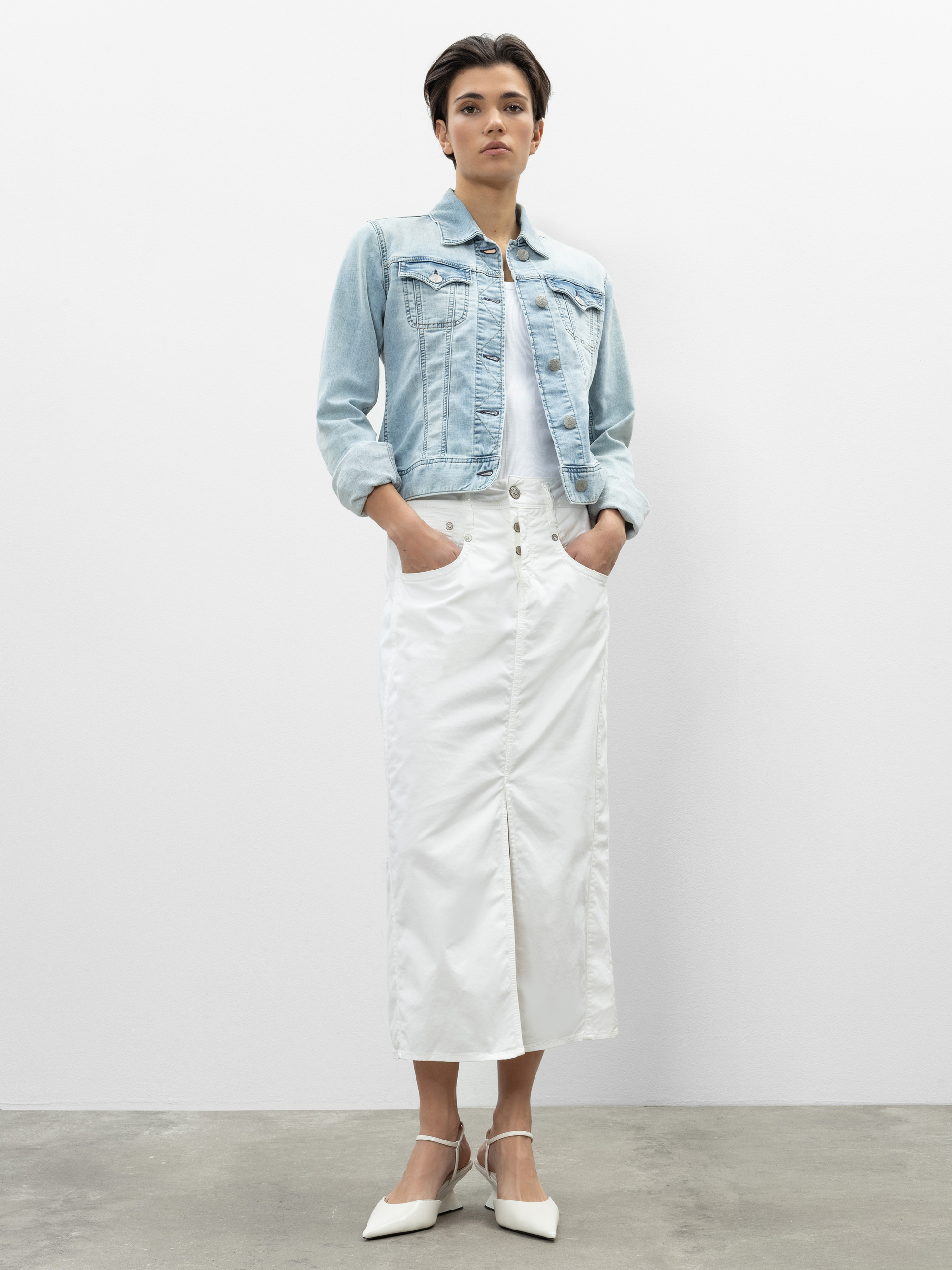 Eine Person steht vor einer schlichten weißen Wand, trägt eine helle Jeansjacke über einem weißen Oberteil, einen langen weißen geknöpften Rock und weiße spitze Absätze, die Hände in den Rocktaschen.