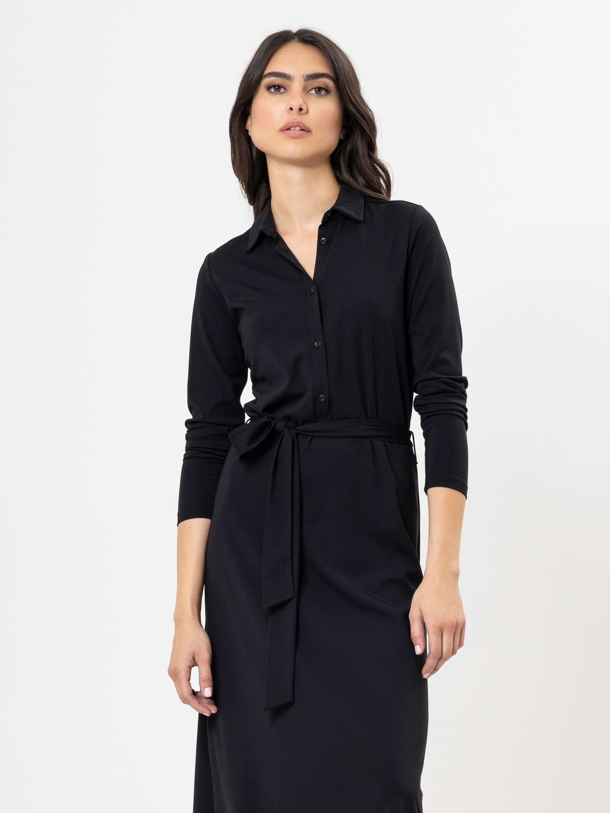 Eine Frau mit langen dunklen Haaren trägt ein schwarzes, langärmeliges, geknöpftes Kleid mit gebundener Taille und steht vor einem schlichten weißen Hintergrund.