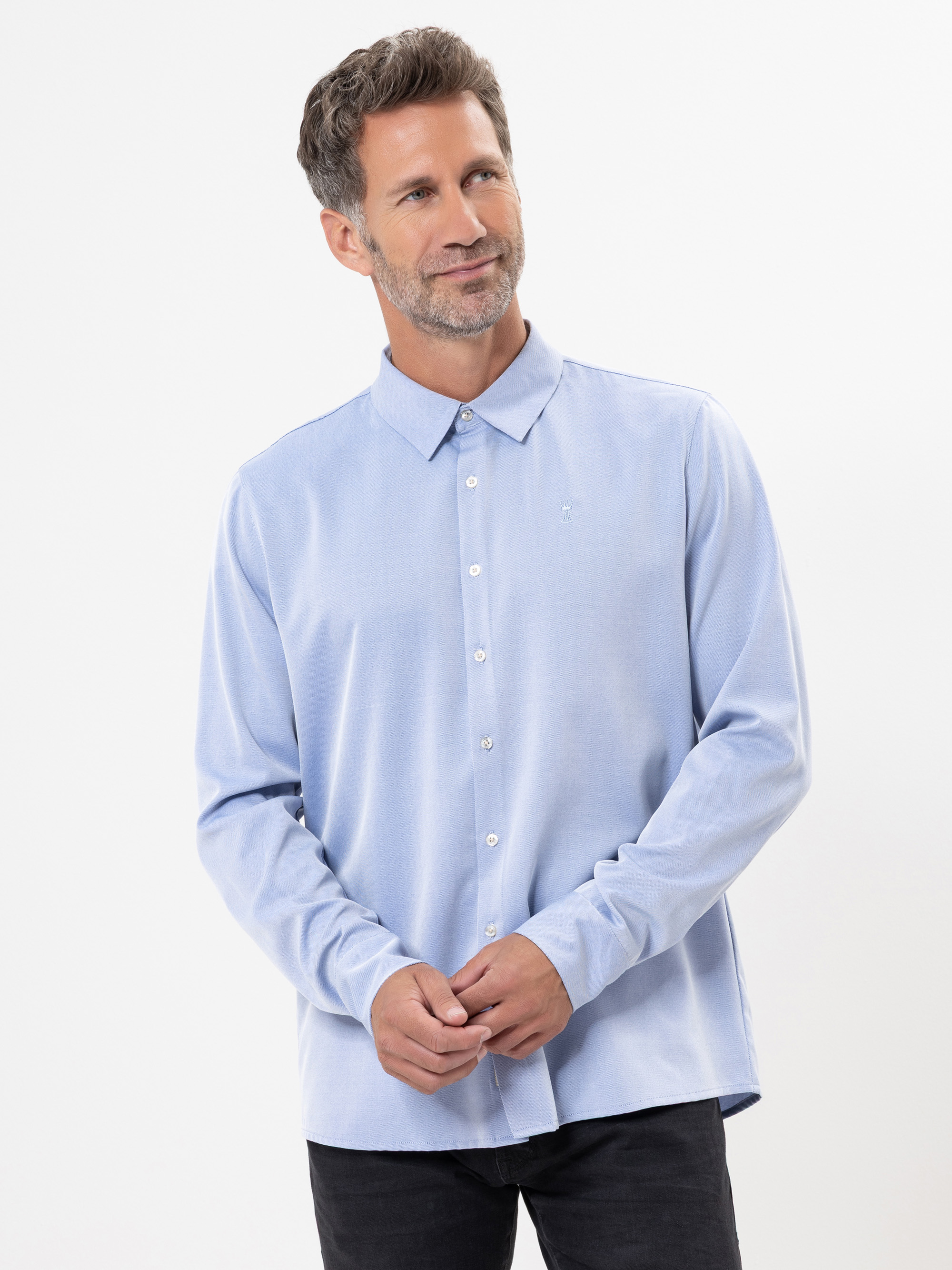 Ein Mann mit kurzen, ergrauten Haaren und einem Bart trägt ein hellblaues Button-up-Hemd und eine schwarze Hose, steht vor einem schlichten weißen Hintergrund und blickt leicht lächelnd zur Seite.