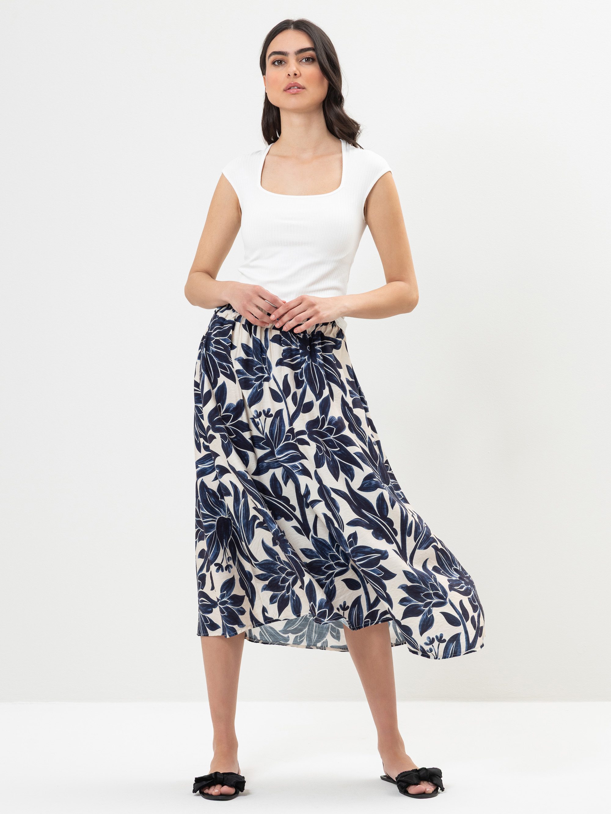 Eine Frau steht vor einem weißen Hintergrund und trägt ein weißes Oberteil mit langen Ärmeln und einen fließenden, knöchellangen Rock mit einem marineblauen Blumenmuster. Sie hat langes dunkles Haar und schwarze Slide-Sandalen.