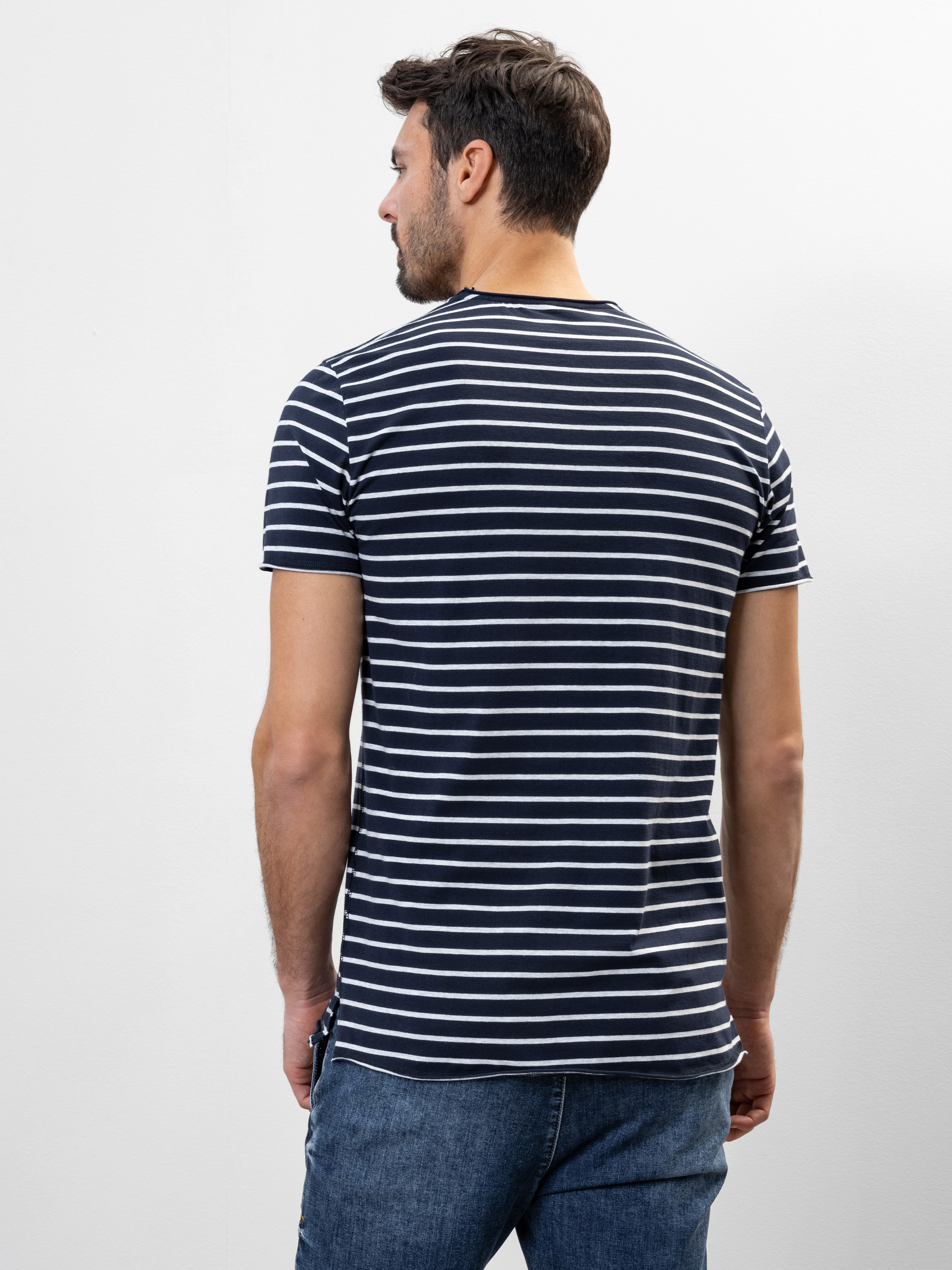 Ein Mann steht mit dem Rücken zur Kamera, trägt ein schwarz-weißes, horizontal gestreiftes T-Shirt und blaue Jeans vor einem schlichten, hellen Hintergrund.