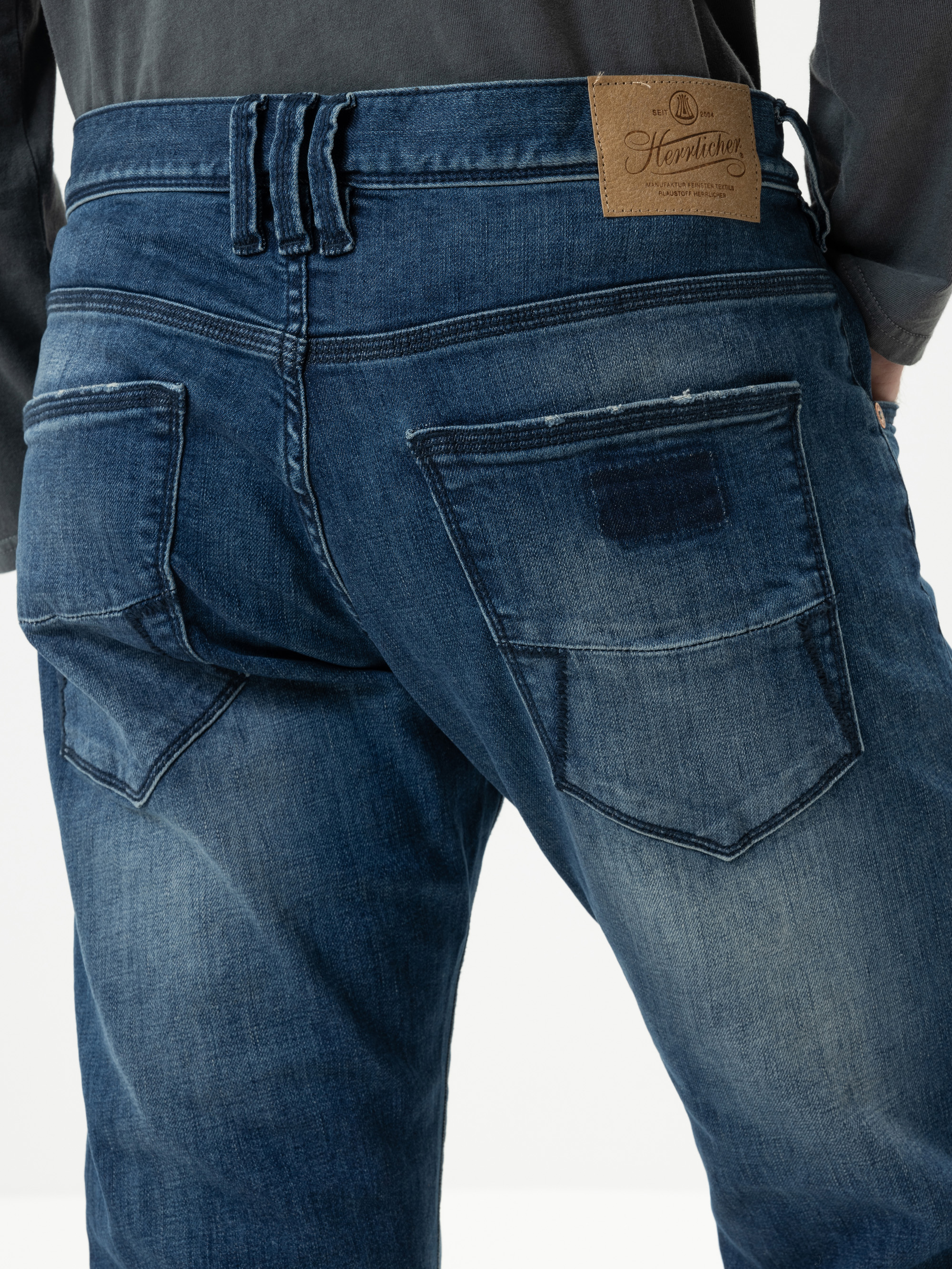 Nahaufnahme des Rückens einer Person, die verblichene blaue Jeans mit einem braunen Lederaufnäher am Bund und großen Gesäßtaschen trägt und vor einem schlichten Hintergrund steht.