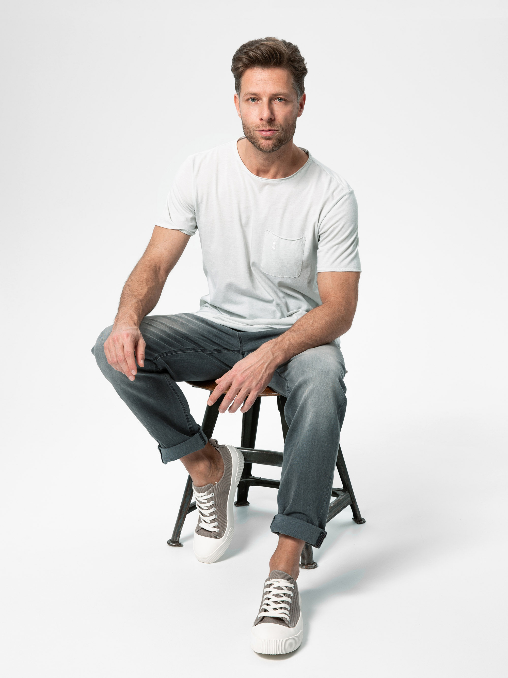 Ein Mann mit kurzen braunen Haaren und einem Bart sitzt auf einem Hocker vor einem weißen Hintergrund. Er trägt ein hellgraues T-Shirt, dunkelgraue, hochgekrempelte Jeans und graue Turnschuhe mit weißen Sohlen.