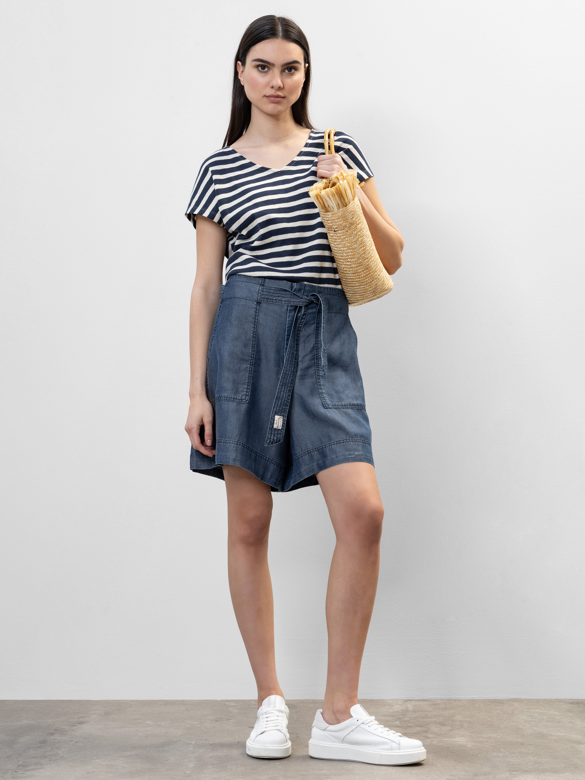 Eine Frau steht vor einer schlichten weißen Wand und trägt ein schwarz-weiß gestreiftes T-Shirt, einen blauen Wickelrock und weiße Turnschuhe. Sie trägt eine geflochtene Strohtasche und hat lange dunkle Haare.