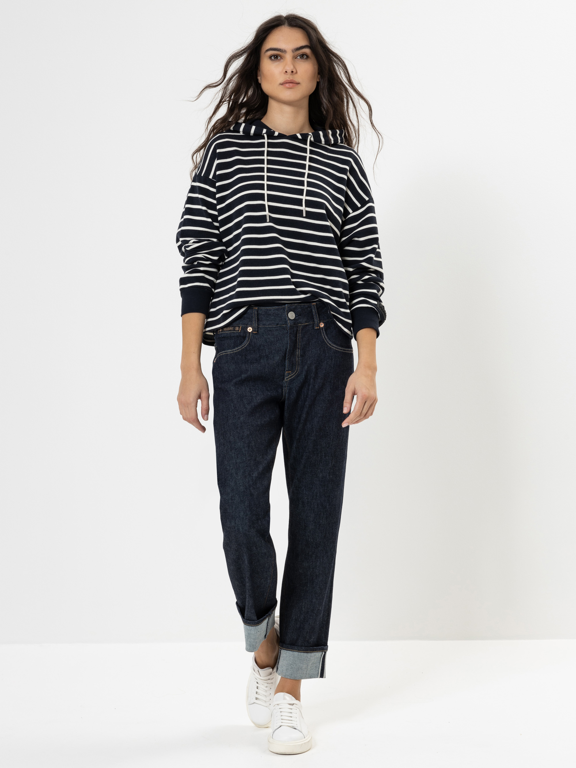 Eine Person steht vor einem schlichten weißen Hintergrund und trägt einen schwarz-weiß gestreiften Kapuzenpullover, dunkle Jeans mit Bündchen und weiße Turnschuhe. Ihr langes Haar liegt offen, und sie hat einen neutralen Gesichtsausdruck.