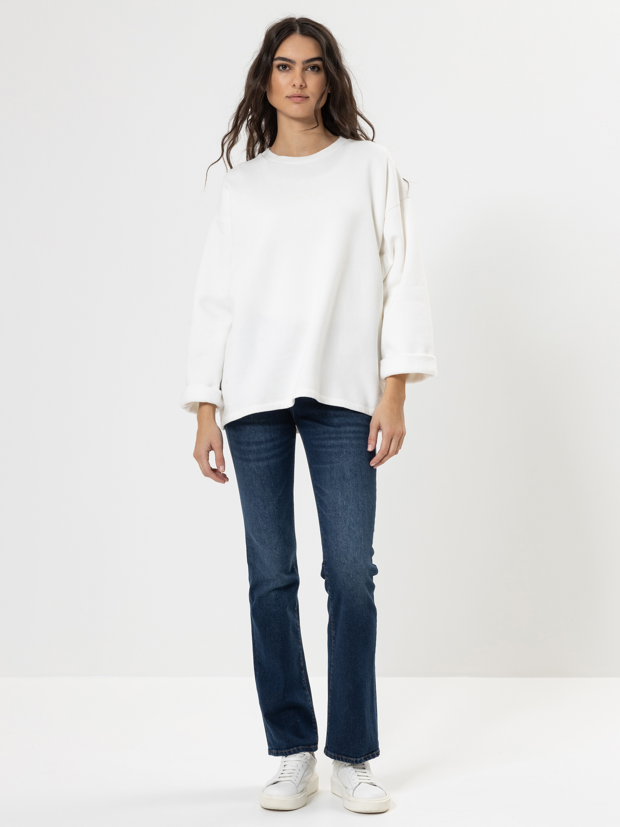 Eine Person steht vor einem schlichten weißen Hintergrund und trägt ein lockeres weißes Sweatshirt, dunkelblaue Jeans und weiße Turnschuhe. Sie hat langes, gewelltes Haar und einen neutralen Gesichtsausdruck, die Arme liegen entspannt an der Seite.