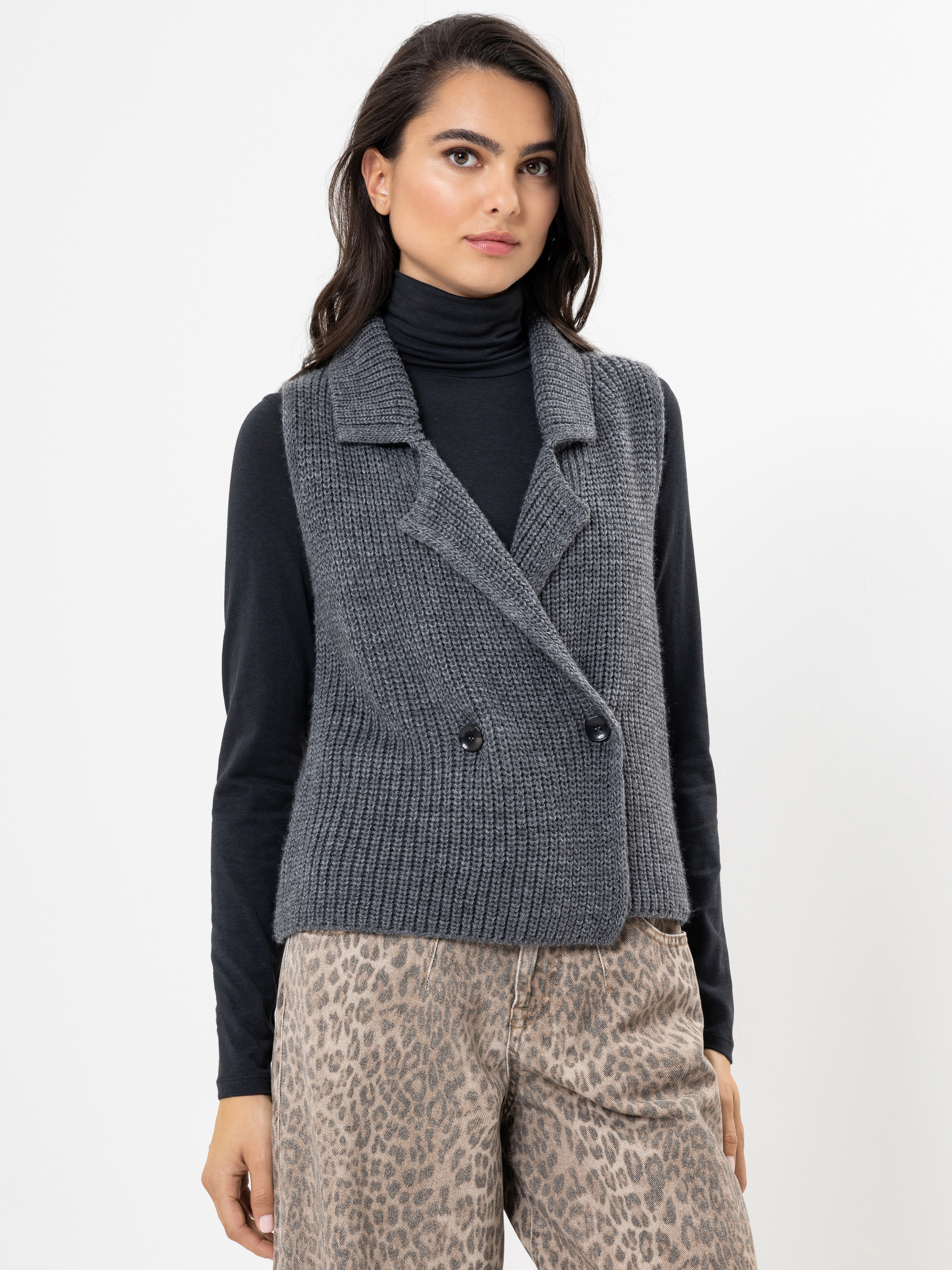 Eine Frau, die einen dunklen Rollkragenpullover, eine graue, doppelreihige Strickweste und eine hochtaillierte, hellbraune Hose mit Tiermuster trägt, steht vor einem schlichten weißen Hintergrund.