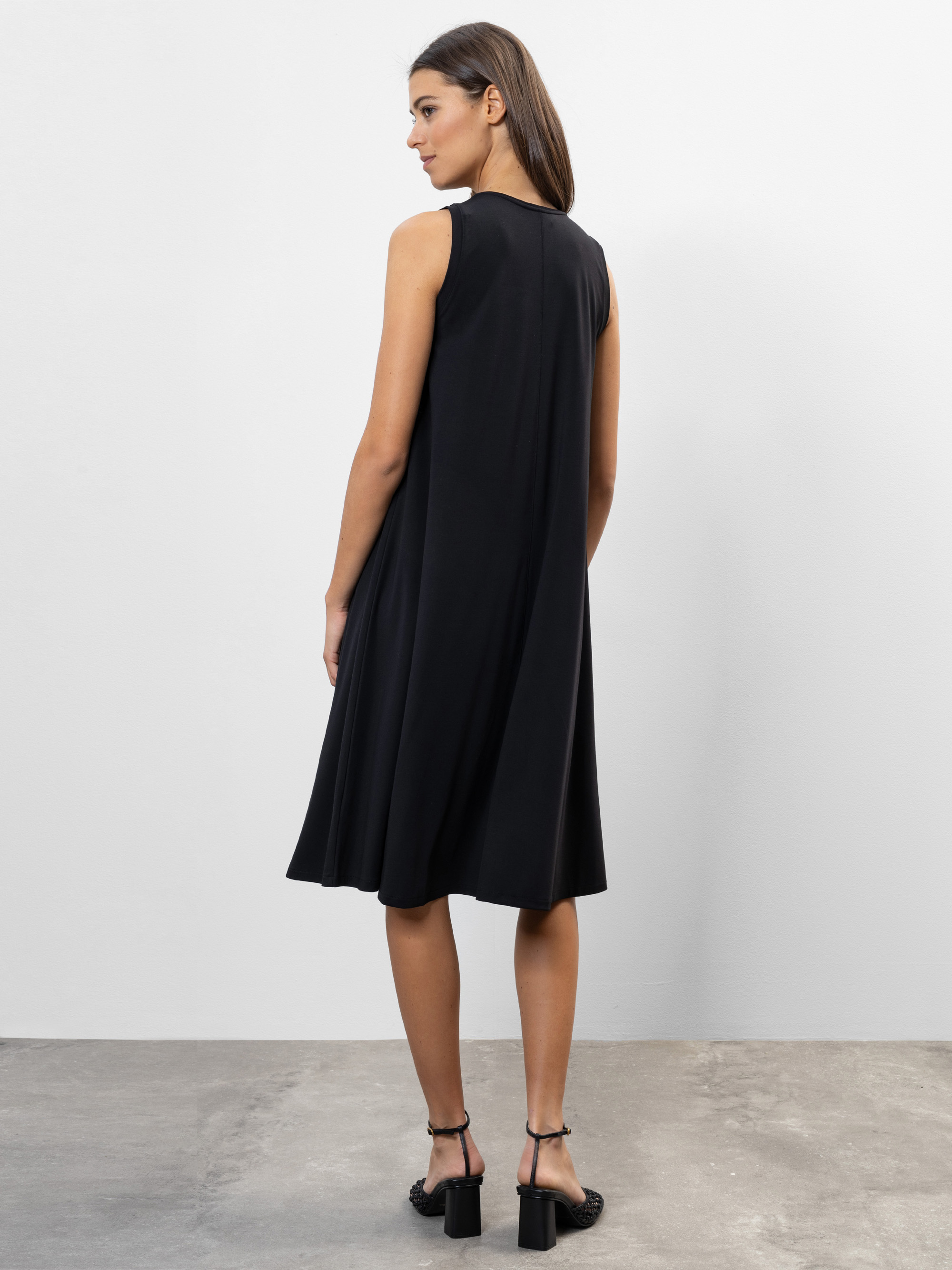 Eine Frau mit glattem braunem Haar trägt ein ärmelloses, knielanges schwarzes Kleid und schwarze Sandaletten mit Absatz. Sie steht auf einem Betonboden und blickt von der Kamera weg in einen minimalistischen, weißwandigen Raum.