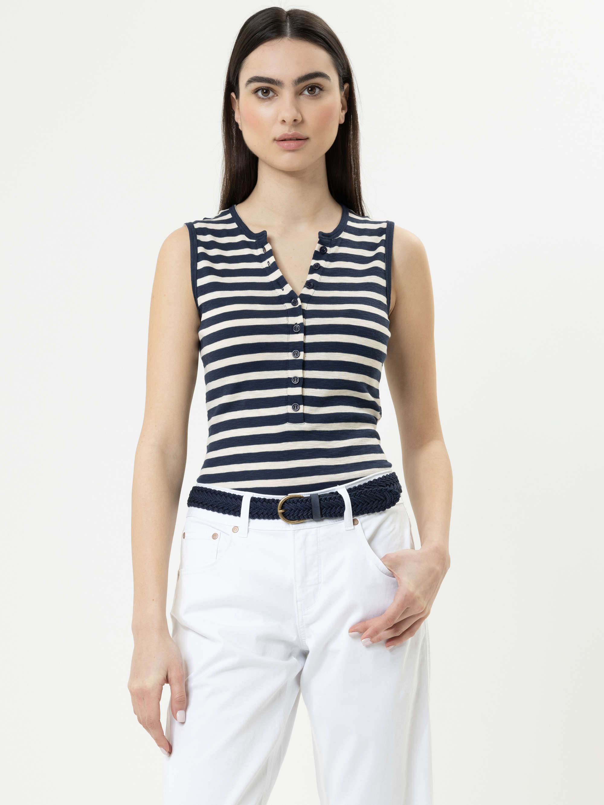 Eine Frau mit glattem dunklem Haar trägt ein marineblau-weiß gestreiftes ärmelloses Oberteil mit Knöpfen und eine weiße Hose. Sie steht vor einem schlichten, hellen Hintergrund und hat eine Hand in ihrer Tasche.