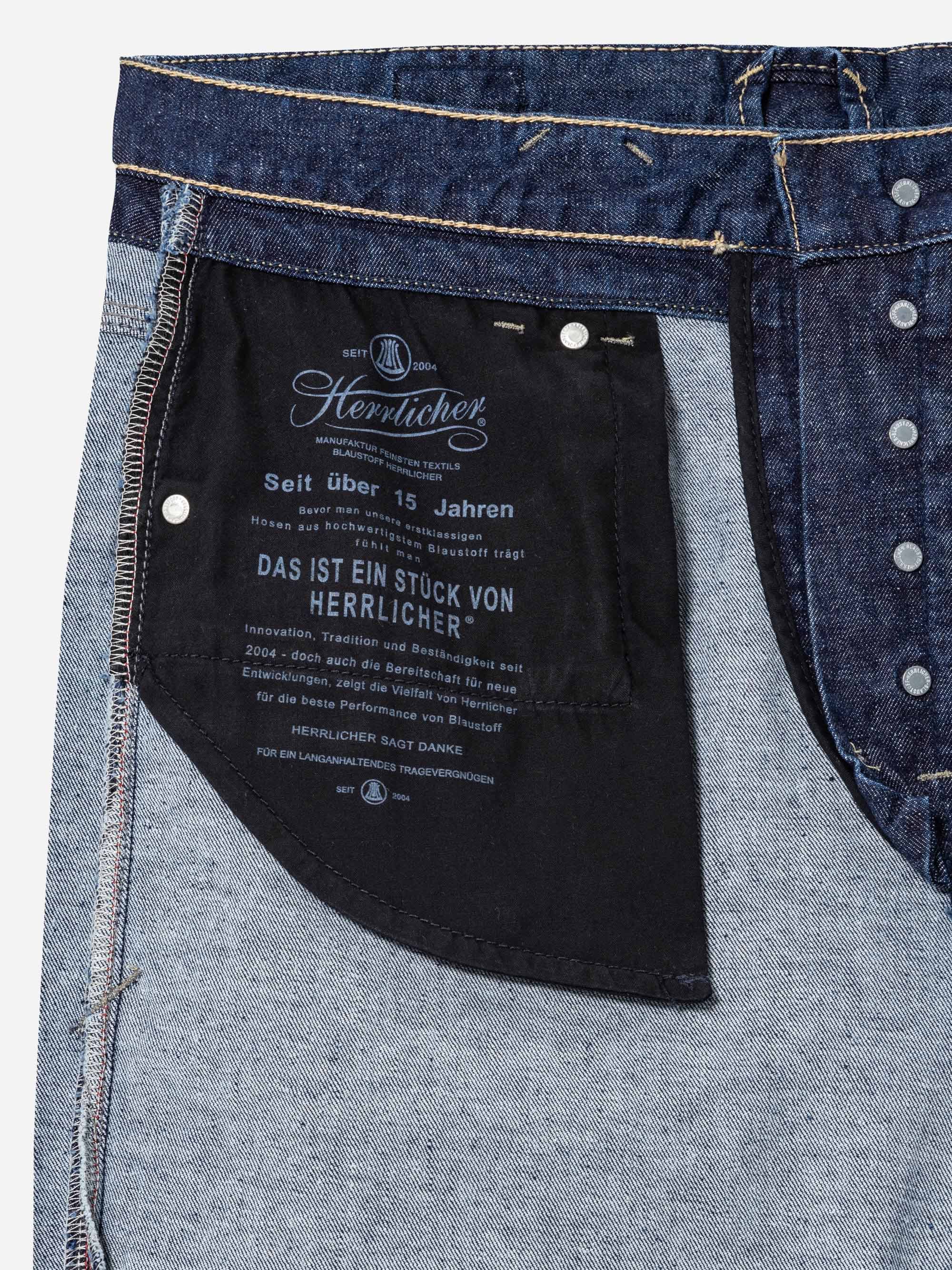 Nahaufnahme der Innenseite einer Jeans, die das Taschenfutter mit aufgedrucktem Branding und Text in deutscher Sprache, Metallnieten, Reißverschluss und kontrastierende Jeansstoffe zeigt.