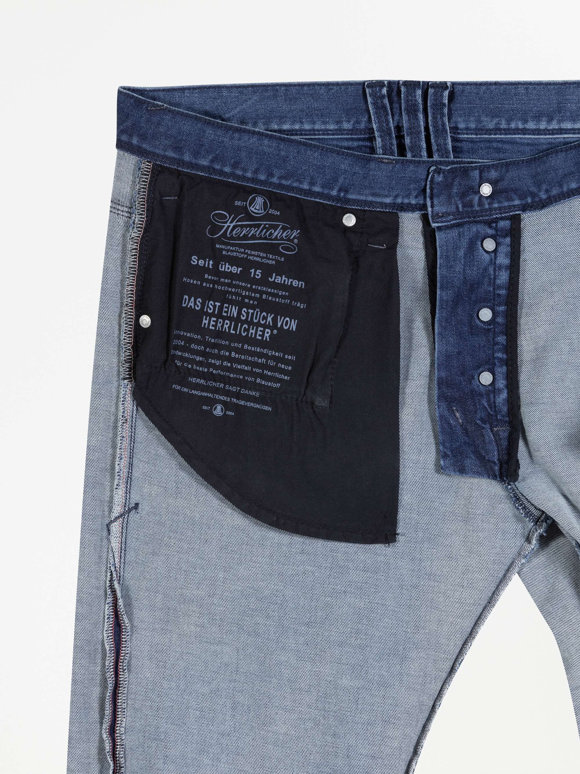 Eine umgedrehte Jeans, die den Bund, den Hosenschlitz mit Knöpfen, das Futter der Innentasche und den auf dem schwarzen Taschenstoff aufgedruckten Text in deutscher Sprache zeigt.