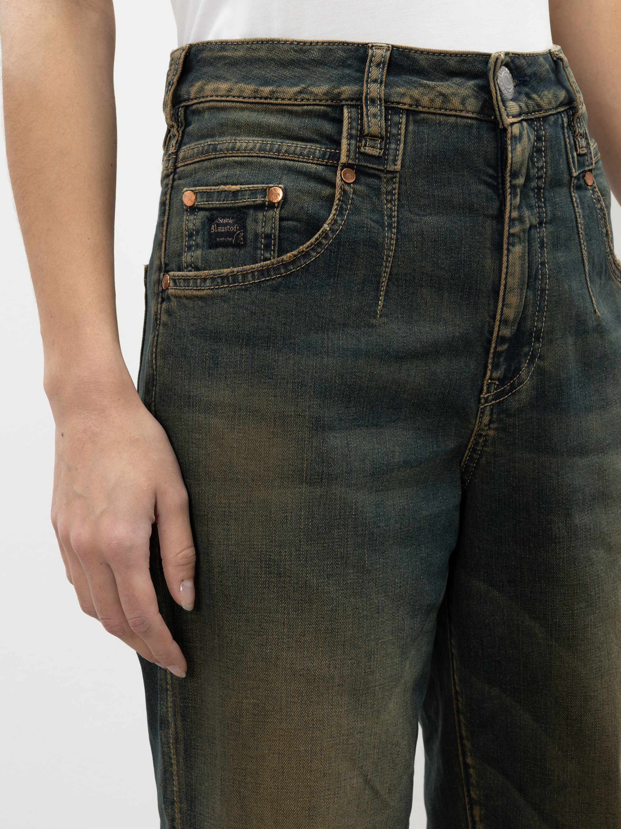 Eine Person trägt eine verblichene blaue High-Waisted-Jeans und ein weißes Hemd, die linke Hand ruht an der Seite. Die Jeans ist mit Kupfernieten und einer kleinen Vordertasche versehen.