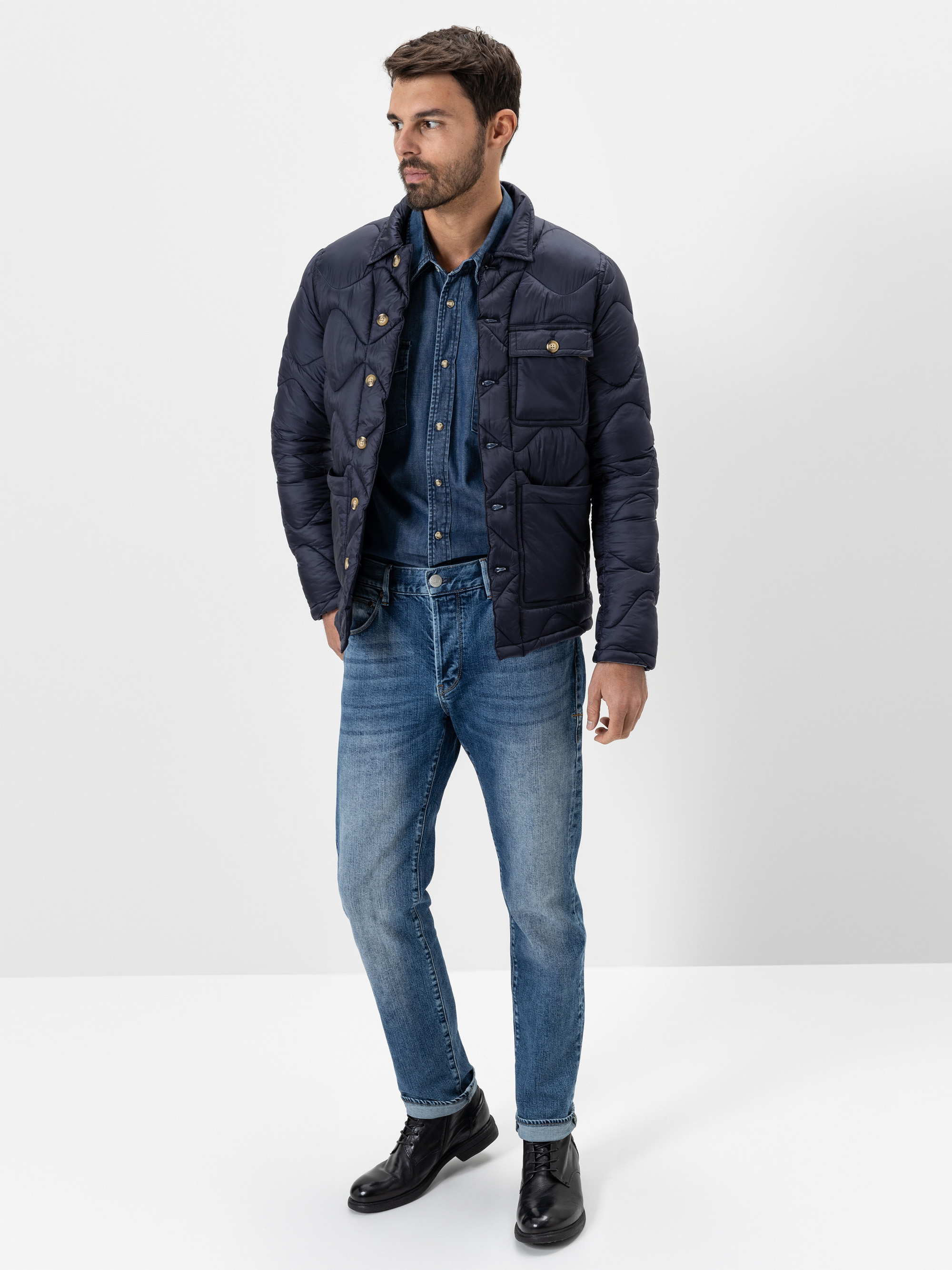 Ein Mann steht vor einem schlichten weißen Hintergrund und trägt eine marineblaue wattierte Jacke über einem Jeanshemd und blaue Jeans, dazu schwarze Schnürstiefel. Er hat kurzes dunkles Haar und einen Bart und blickt zur Seite.