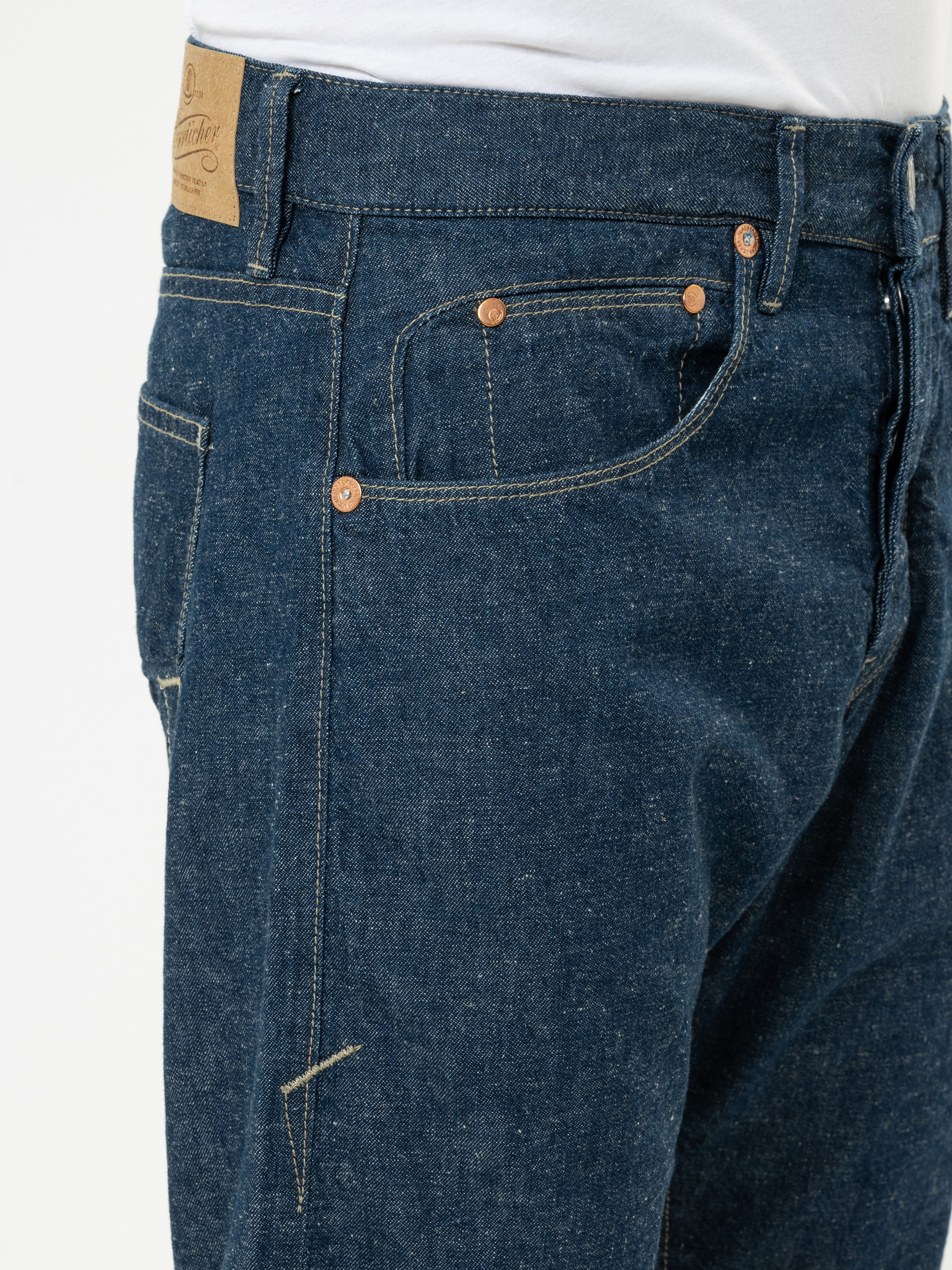 Nahaufnahme einer Person, die eine blaue Jeans mit kupferfarbenen Nieten und hellbraunen Nähten trägt, wobei die Vordertasche, die Münztasche und ein Teil eines braunen Lederflickens am Bund zu sehen sind. Die Person trägt außerdem ein weißes Hemd.