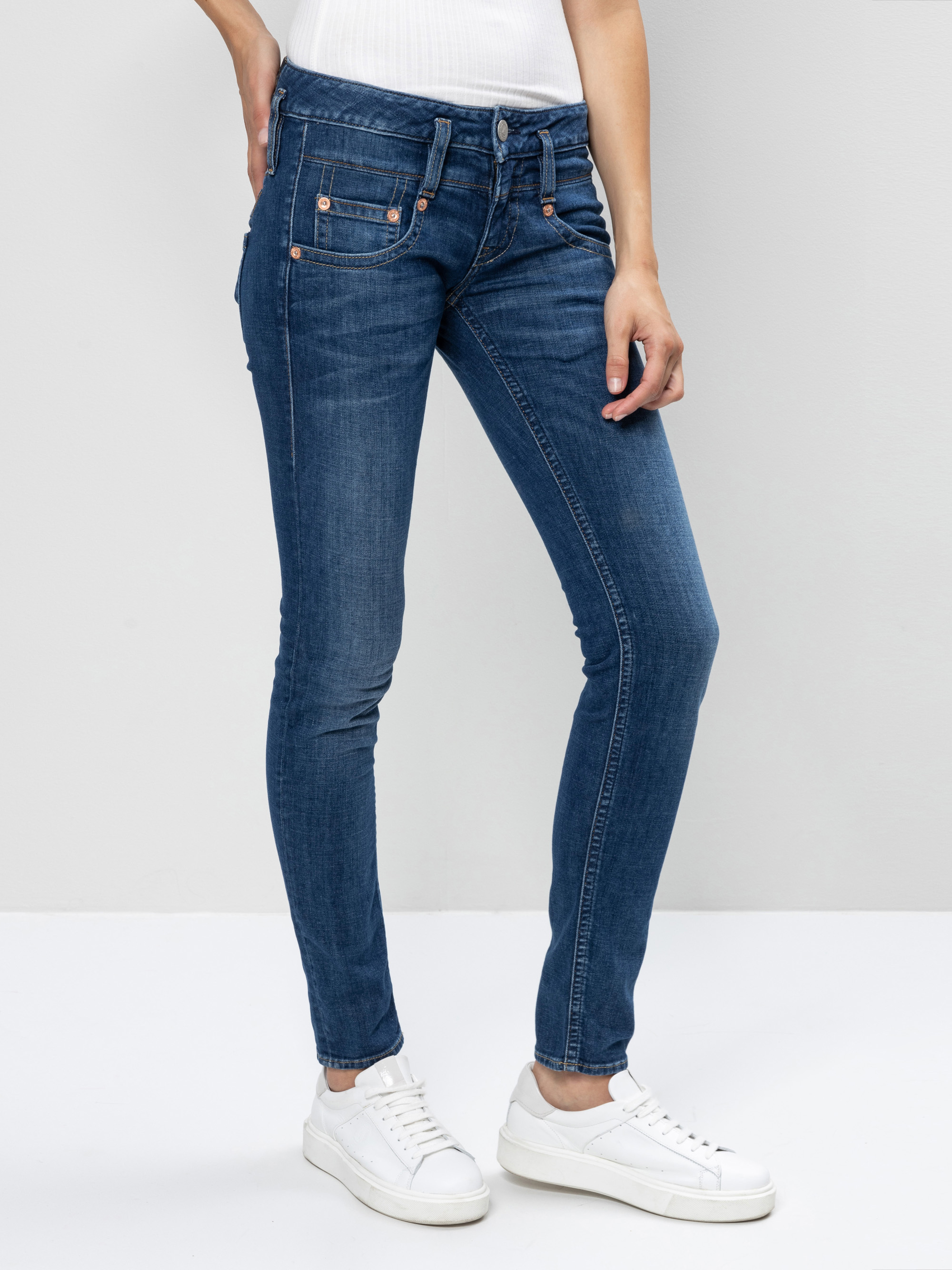 Eine Person, die ein weißes ärmelloses Oberteil, blaue Skinny-Jeans und weiße Turnschuhe trägt, steht vor einem schlichten hellgrauen Hintergrund. Das Bild ist oberhalb der Schultern abgeschnitten.