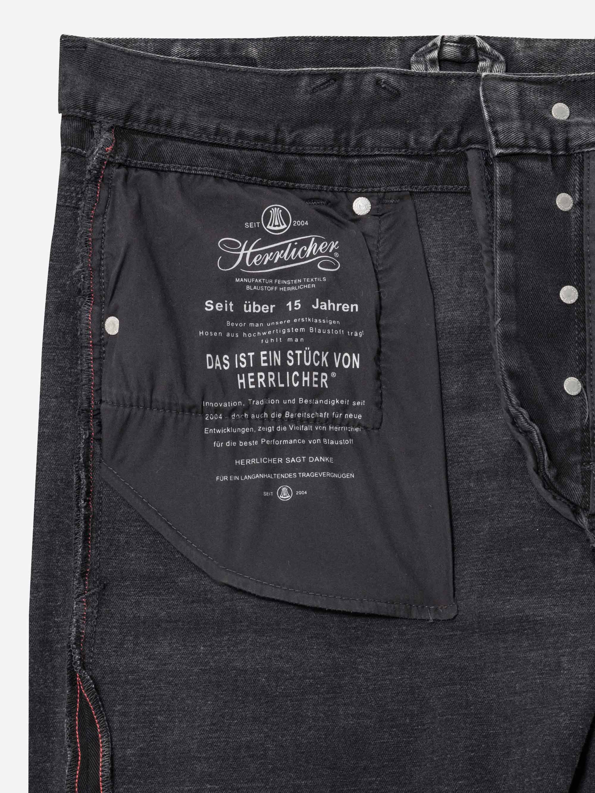 Nahaufnahme der Innentasche einer schwarzen Jeans, auf der ein weißer Text mit Branding und Produktinformationen der Marke Herrlicher aufgedruckt ist. Die Knöpfe und Nahtdetails sind ebenfalls sichtbar.
