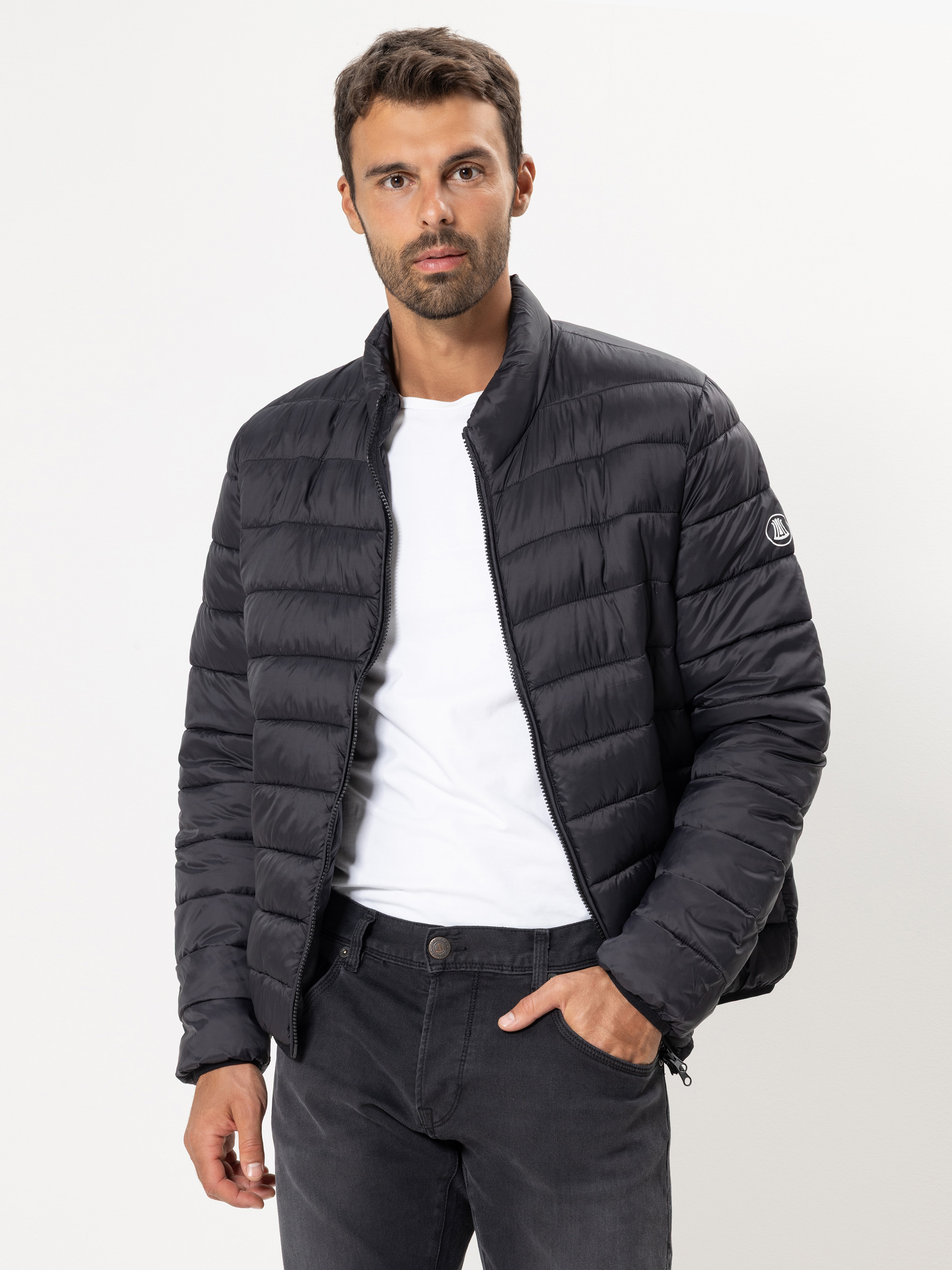 Ein Mann mit kurzen dunklen Haaren und einem Bart trägt eine schwarze Steppjacke über einem weißen T-Shirt und dunkle Jeans. Er steht vor einem schlichten weißen Hintergrund und hat eine Hand in der Tasche.