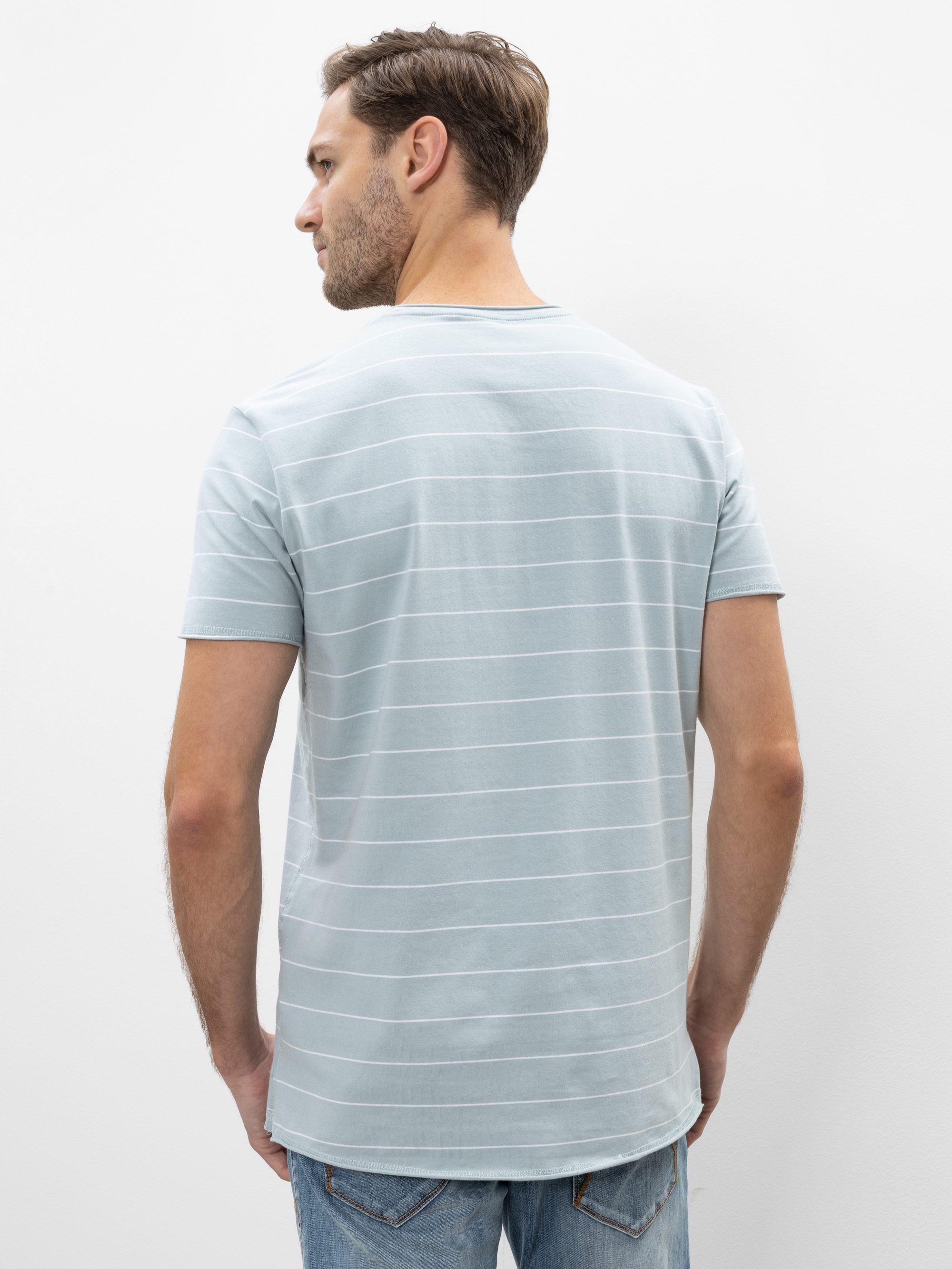 Ein Mann mit kurzen braunen Haaren trägt ein hellblaues, kurzärmeliges, gestreiftes T-Shirt und blaue Jeans und steht mit dem Rücken zur Kamera vor einem schlichten weißen Hintergrund.