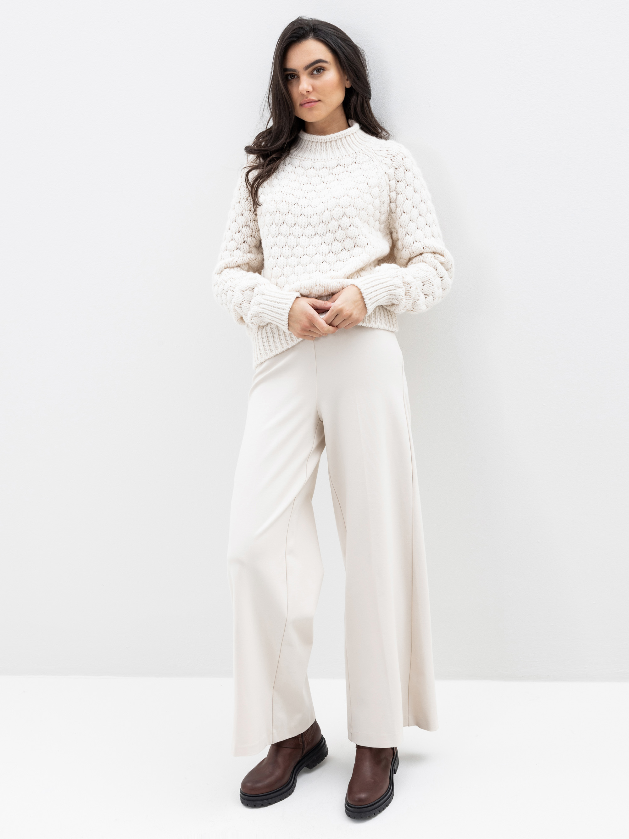 Eine Frau steht vor einer weißen Wand und trägt einen cremefarbenen Pullover mit Struktur, eine cremefarbene Hose mit weitem Bein und dunkelbraune Stiefeletten. Sie hat langes dunkles Haar und einen neutralen Gesichtsausdruck, eine Hand ruht in ihrer Ta