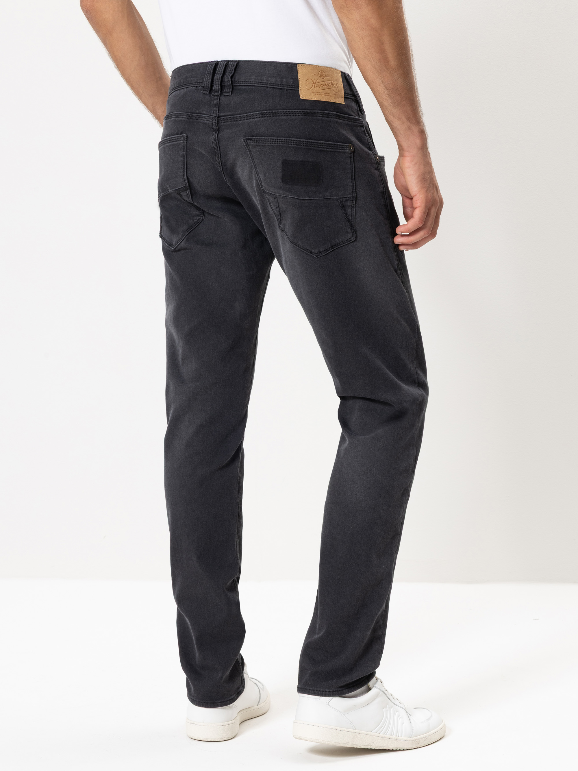 Ein Mann, der schwarze Jeans und weiße Turnschuhe trägt, steht mit dem Rücken zur Kamera vor einem schlichten weißen Hintergrund.