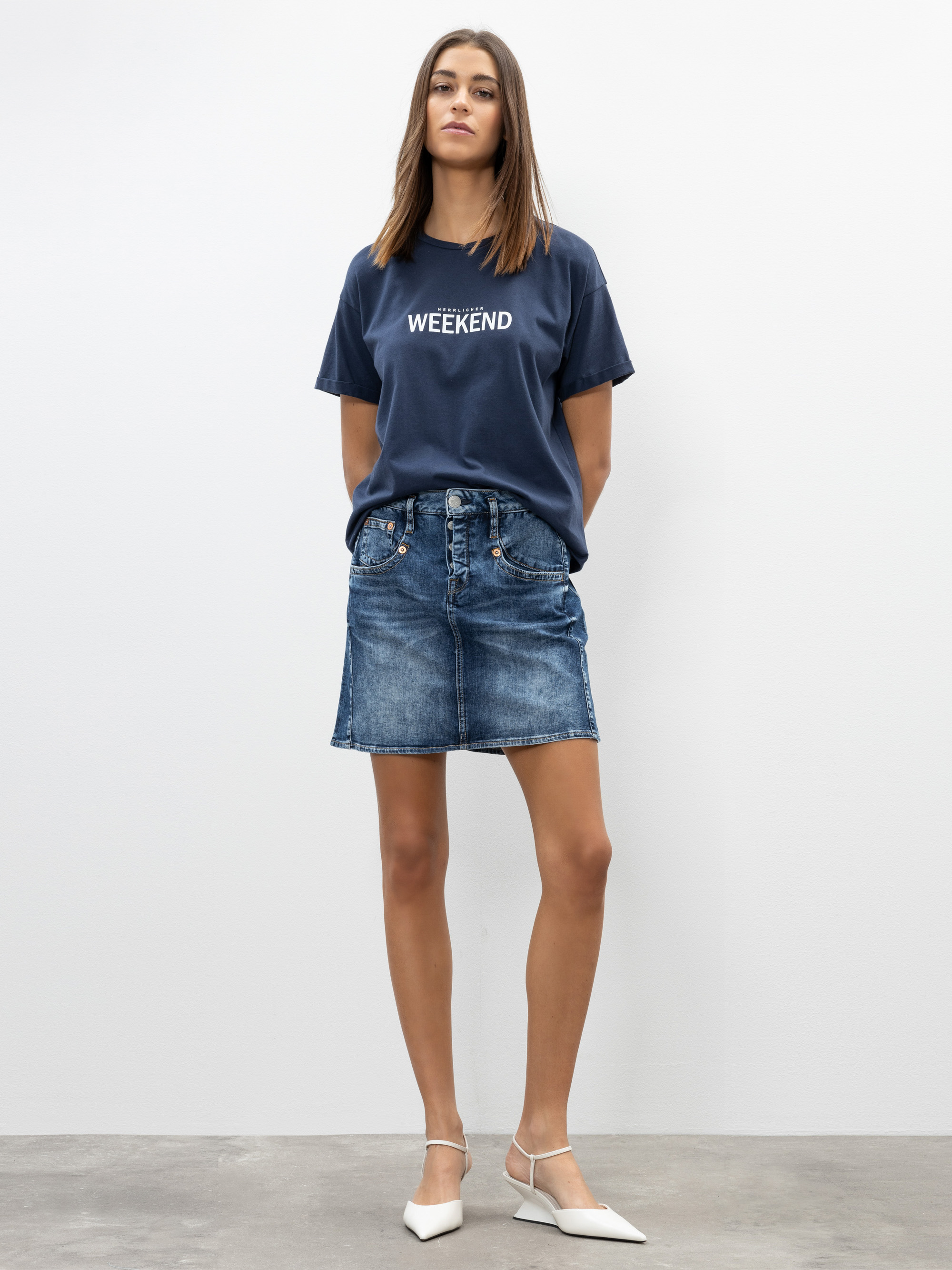 Eine Frau steht vor einer schlichten weißen Wand und trägt ein marineblaues WEEKEND-T-Shirt, einen Jeansminirock und weiße spitze Schuhe mit Knöchelriemen. Ihre Hände stecken in den Rocktaschen und sie schaut selbstbewusst in die Kamera.