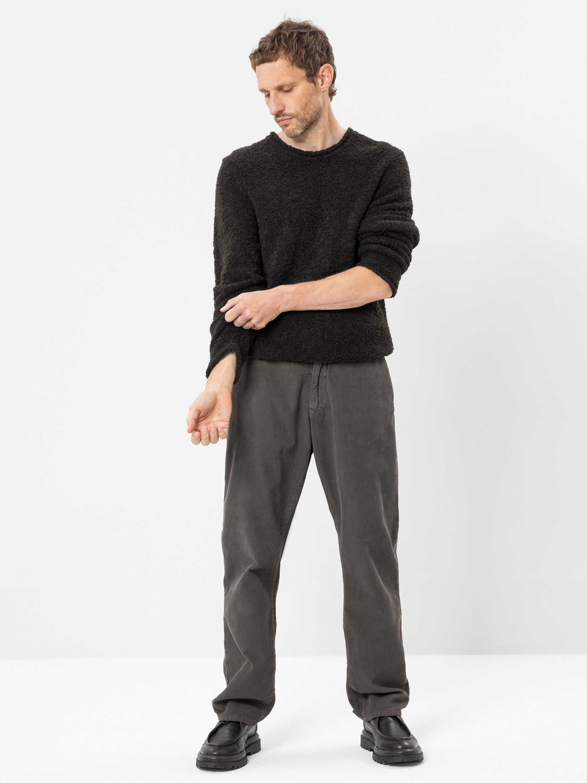 Ein Mann steht vor einem weißen Hintergrund und trägt einen schwarzen Pullover, eine graue Hose und schwarze Schuhe. Er schaut nach unten und krempelt mit der rechten Hand den Ärmel seines linken Arms hoch.