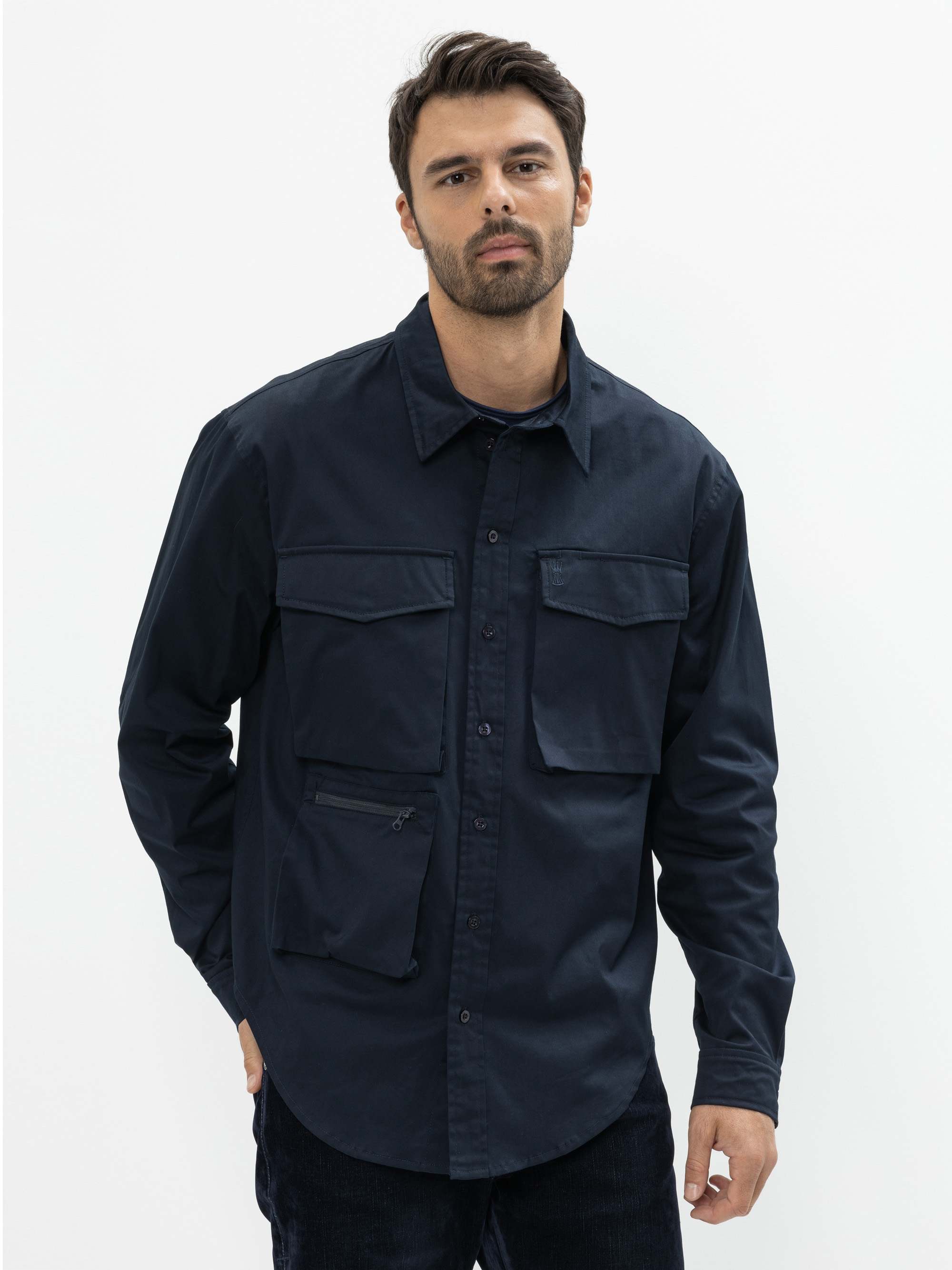 Ein Mann mit kurzen dunklen Haaren und einem Bart trägt ein dunkles, marineblaues Button-up-Hemd mit großen Vordertaschen. Er steht vor einem schlichten weißen Hintergrund und schaut mit neutralem Blick in die Kamera.
