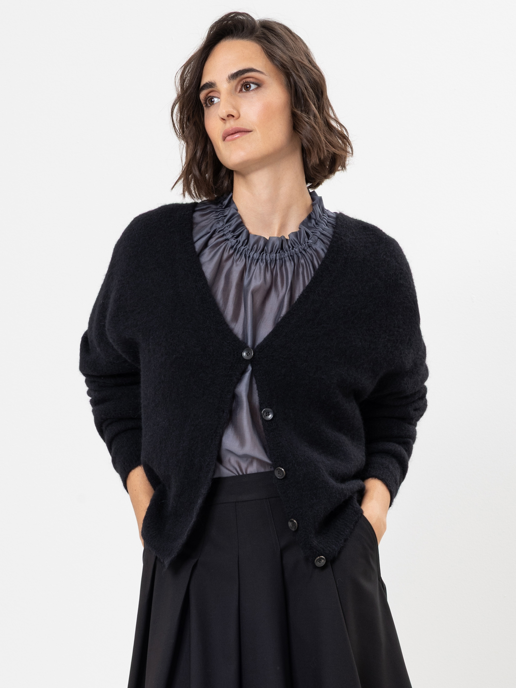 Eine Frau mit kurzen braunen Haaren trägt eine schwarze Strickjacke mit Knöpfen über einer glänzenden grauen Rüschenbluse und einen schwarzen Rock und steht vor einem schlichten weißen Hintergrund. Sie blickt leicht zur Seite und hat die Hände in d