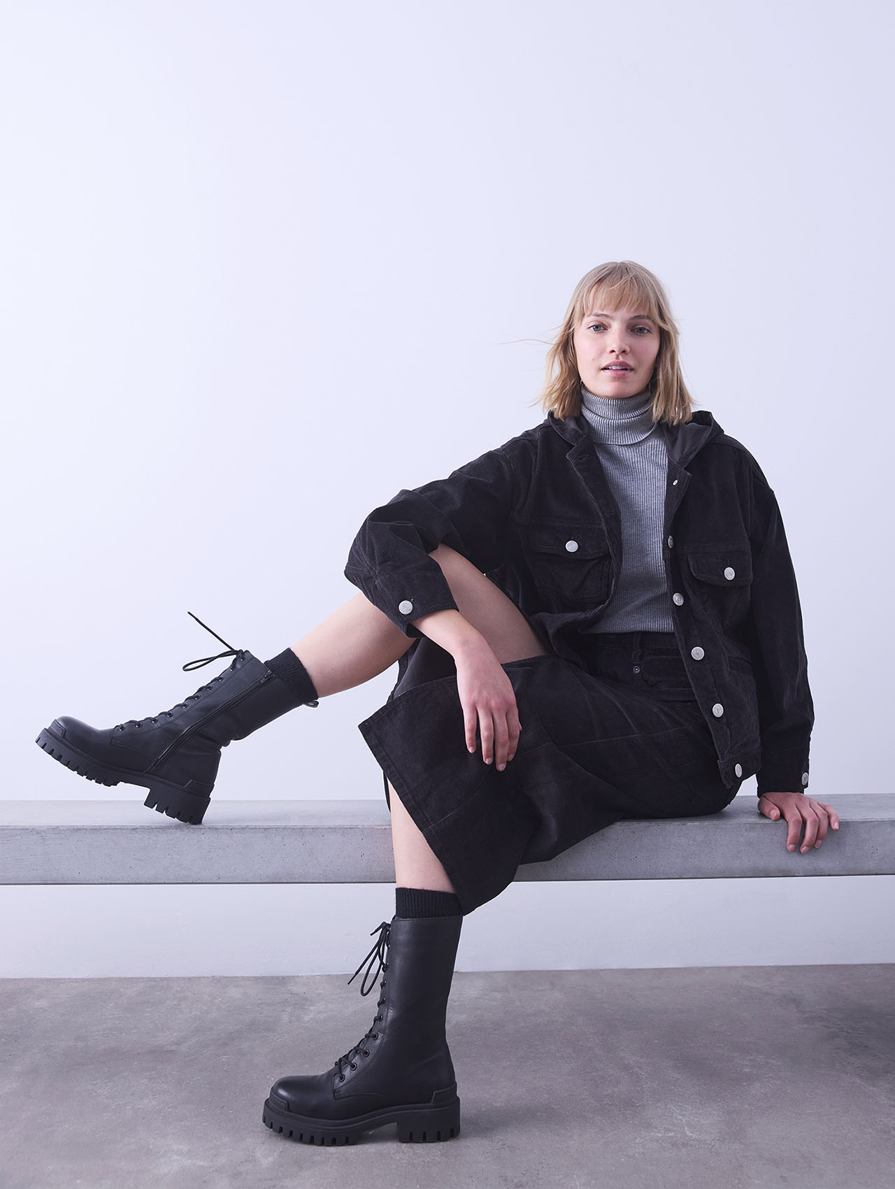 Eine Frau mit blondem Haar sitzt auf einer Bank vor einem schlichten Hintergrund. Sie trägt einen grauen Rollkragenpullover, eine schwarze Jacke und einen Rock sowie schwarze Schnürstiefel. Sie blickt selbstbewusst in die Kamera und stützt ein Bein auf