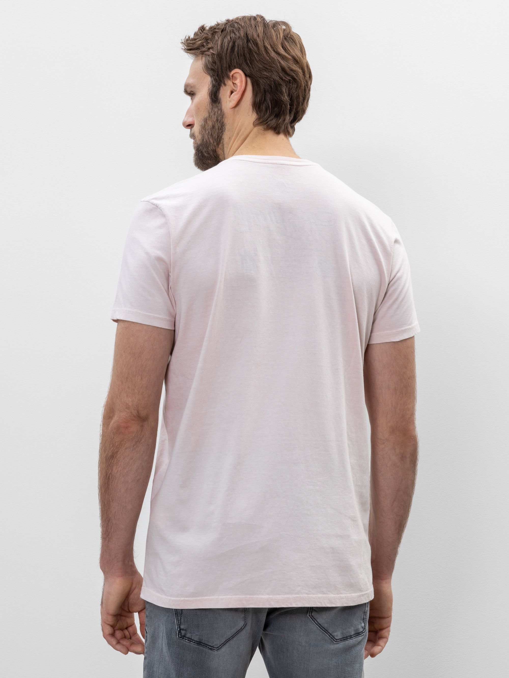 Ein Mann mit hellbraunem Haar und Bart steht mit dem Rücken zur Kamera, trägt ein blassrosa T-Shirt und hellgraue Jeans vor einem schlichten weißen Hintergrund.