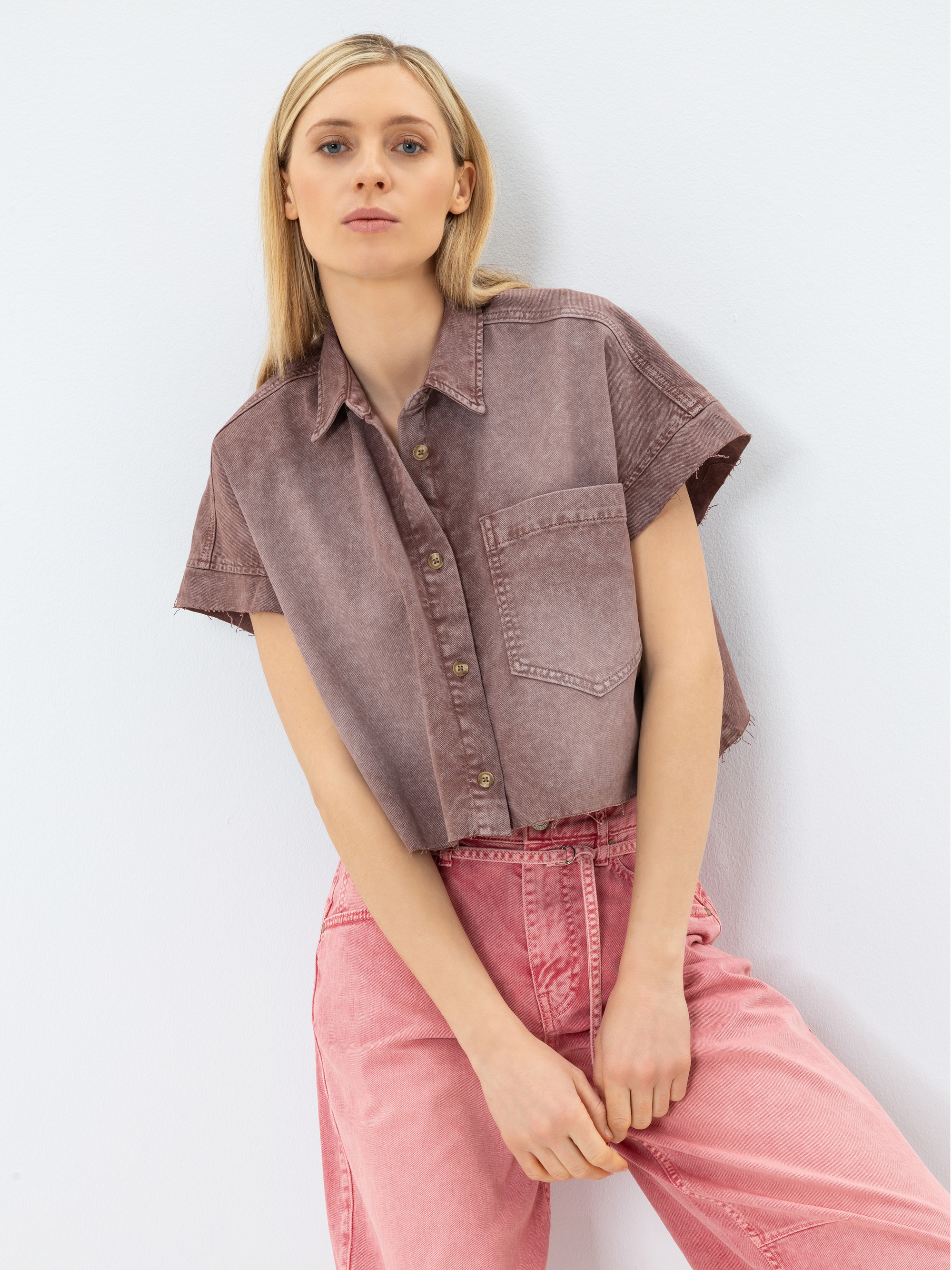 Eine Frau mit glattem blondem Haar trägt ein kurzärmeliges, verblasstes braunes Button-up-Hemd und hochgekrempelte rosa Jeans. Sie steht vor einer weißen Wand und hat die Hände vor sich aufgestützt.