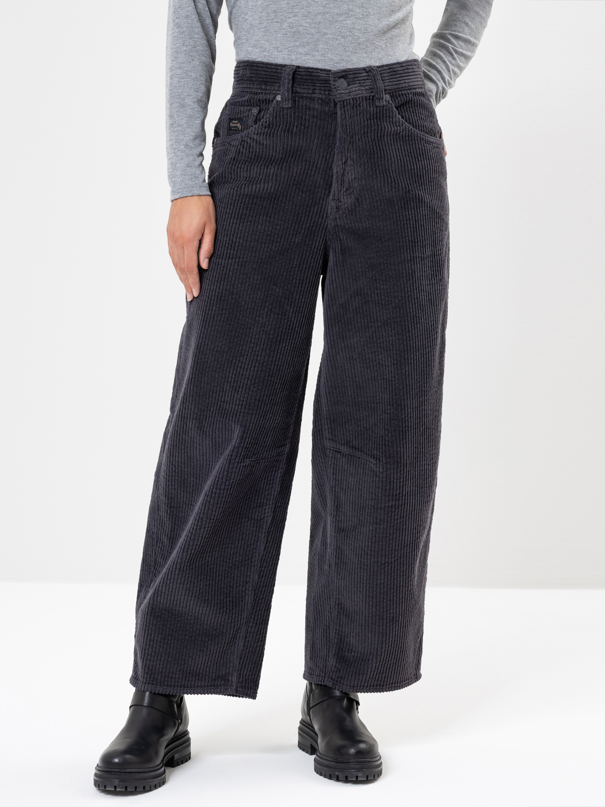 Person, die ein graues langärmeliges Oberteil, eine weite dunkelgraue Cordhose und schwarze Stiefel trägt und vor einem schlichten weißen Hintergrund steht. Das Gesicht der Person ist nicht sichtbar.