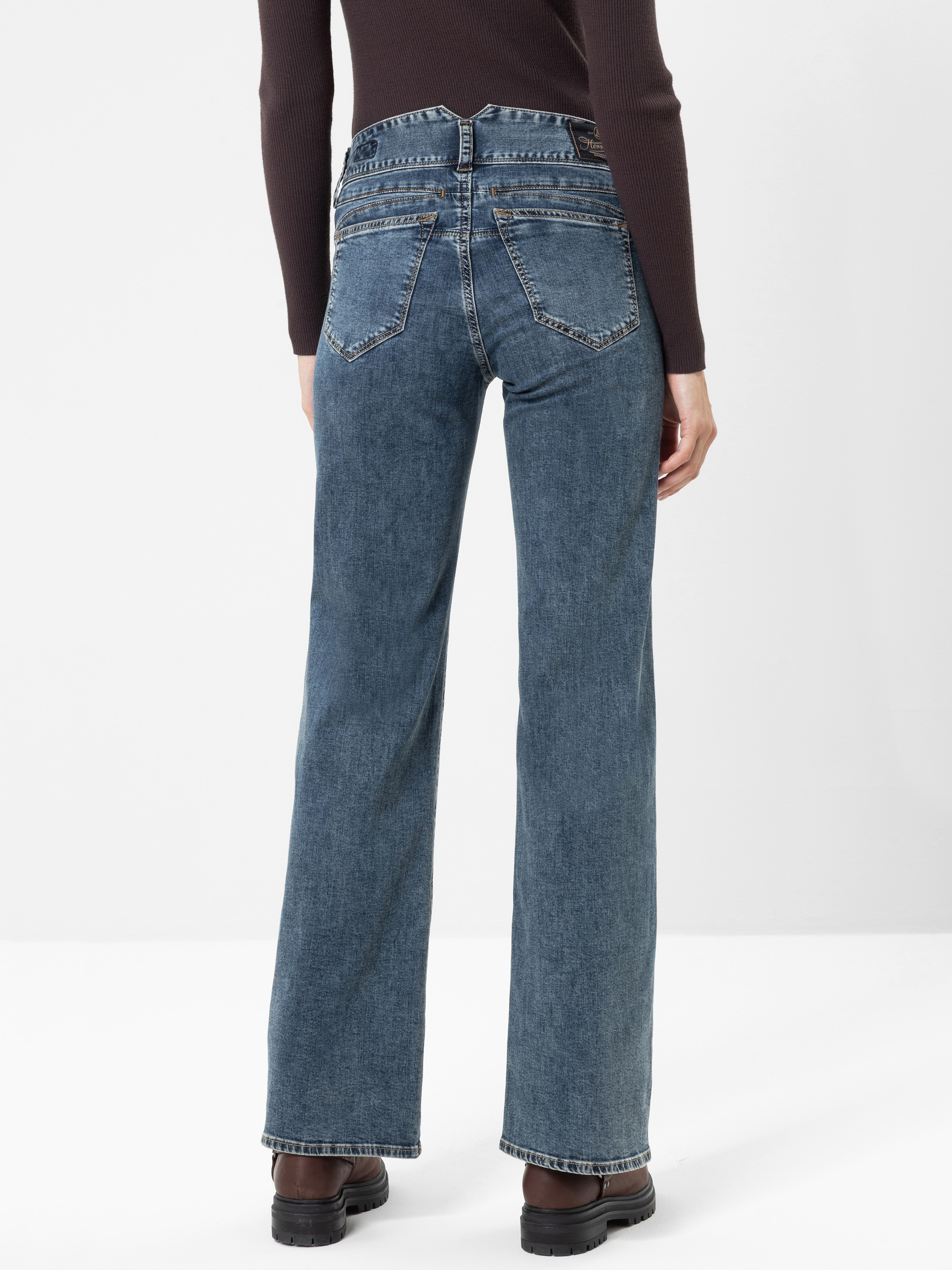 Eine Person, die eine blaue, hoch taillierte Jeans mit geradem Bein, ein dunkles Langarm-Oberteil und schwarze Schuhe trägt, von hinten vor einem schlichten weißen Hintergrund.
