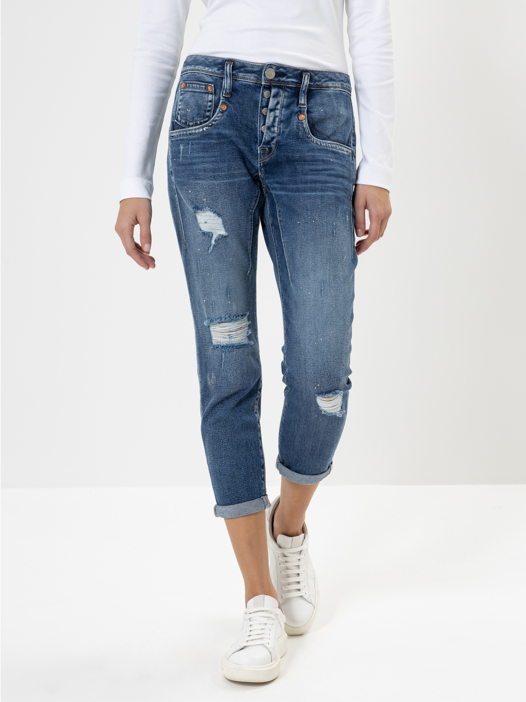 Eine Person, die ein weißes langärmeliges Oberteil, eine Jeans mit aufgerollten Manschetten und weiße Turnschuhe trägt, steht vor einem schlichten weißen Hintergrund.