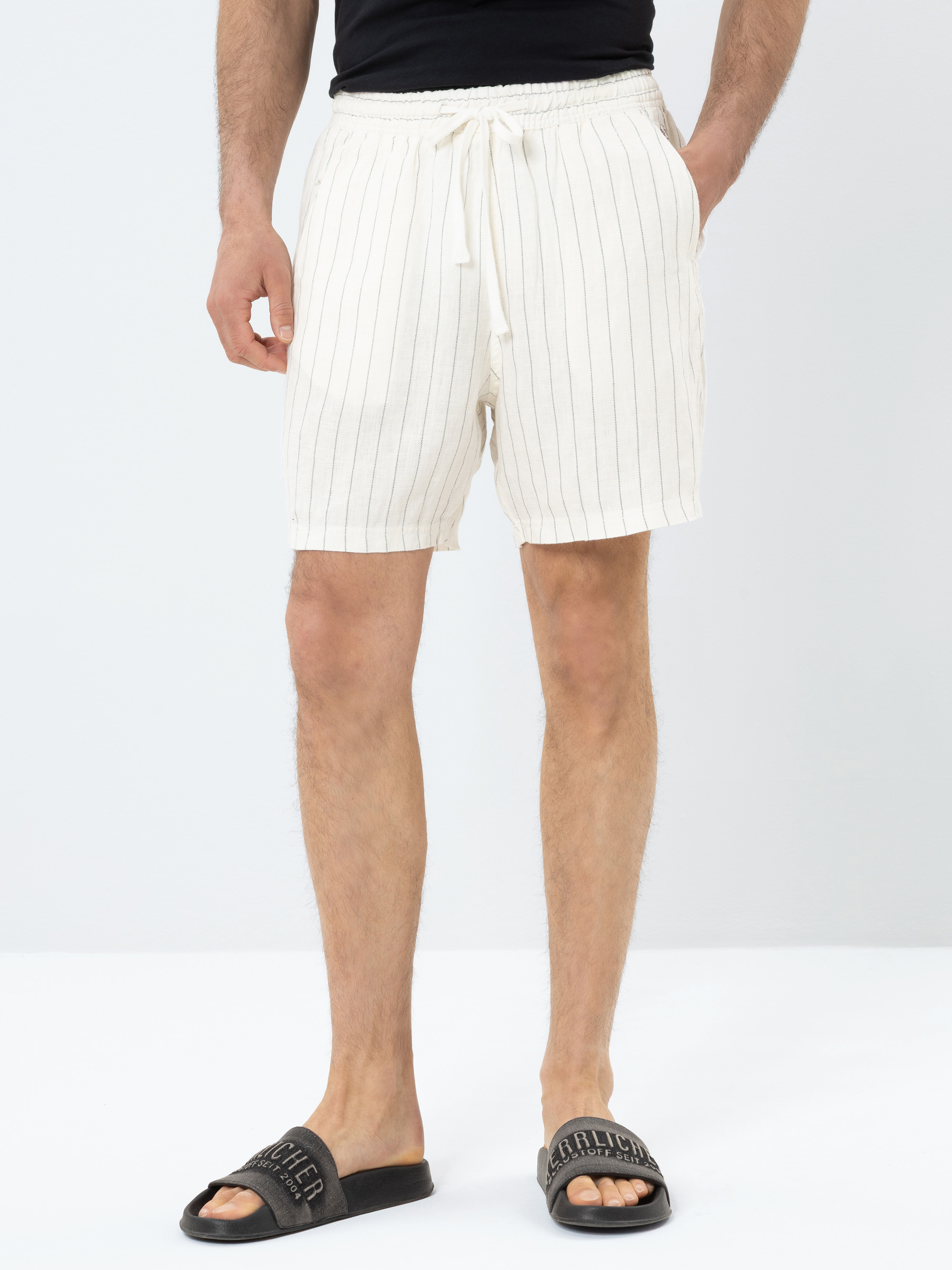Ein Mann, der ein schwarzes Hemd, weiße Shorts mit dünnen vertikalen Streifen und schwarze Slide-Sandalen trägt, steht vor einem schlichten weißen Hintergrund. Seine Hände stecken in den Taschen seiner Shorts.