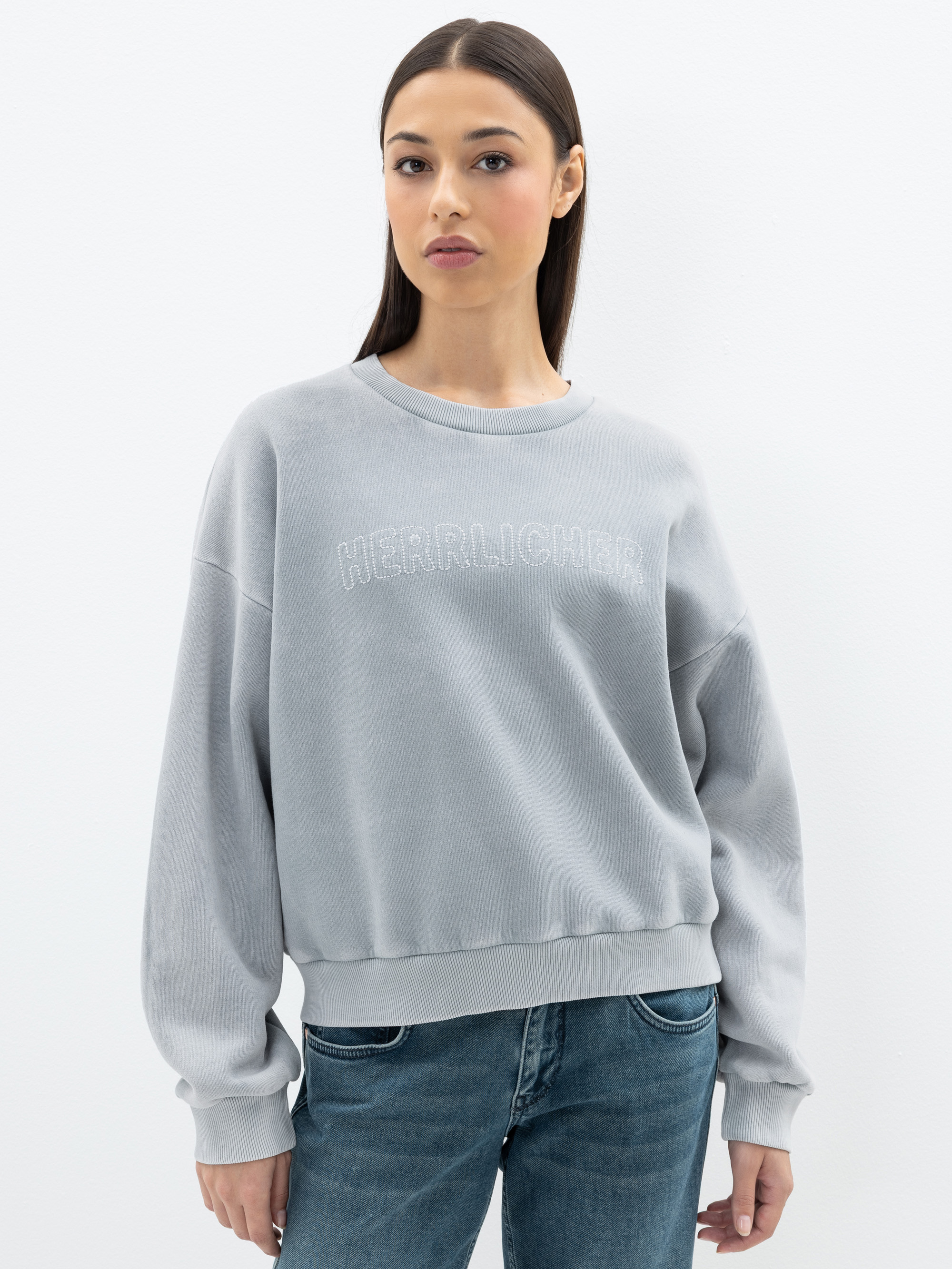 Eine Frau mit glattem dunklem Haar, die ein hellgraues Sweatshirt mit dem aufgestickten Wort HERRLICHER auf der Vorderseite und eine blaue Jeans trägt, steht vor einem schlichten weißen Hintergrund.