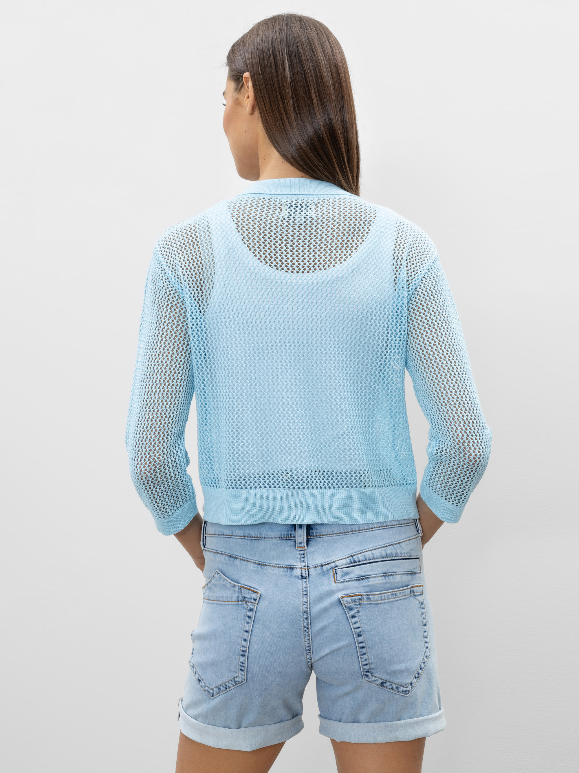 Eine Frau mit glattem braunem Haar trägt einen hellblauen Netzpullover über einem weißen Oberteil und hellblaue Jeansshorts. Sie blickt von der Kamera weg und steht vor einem schlichten hellgrauen Hintergrund.
