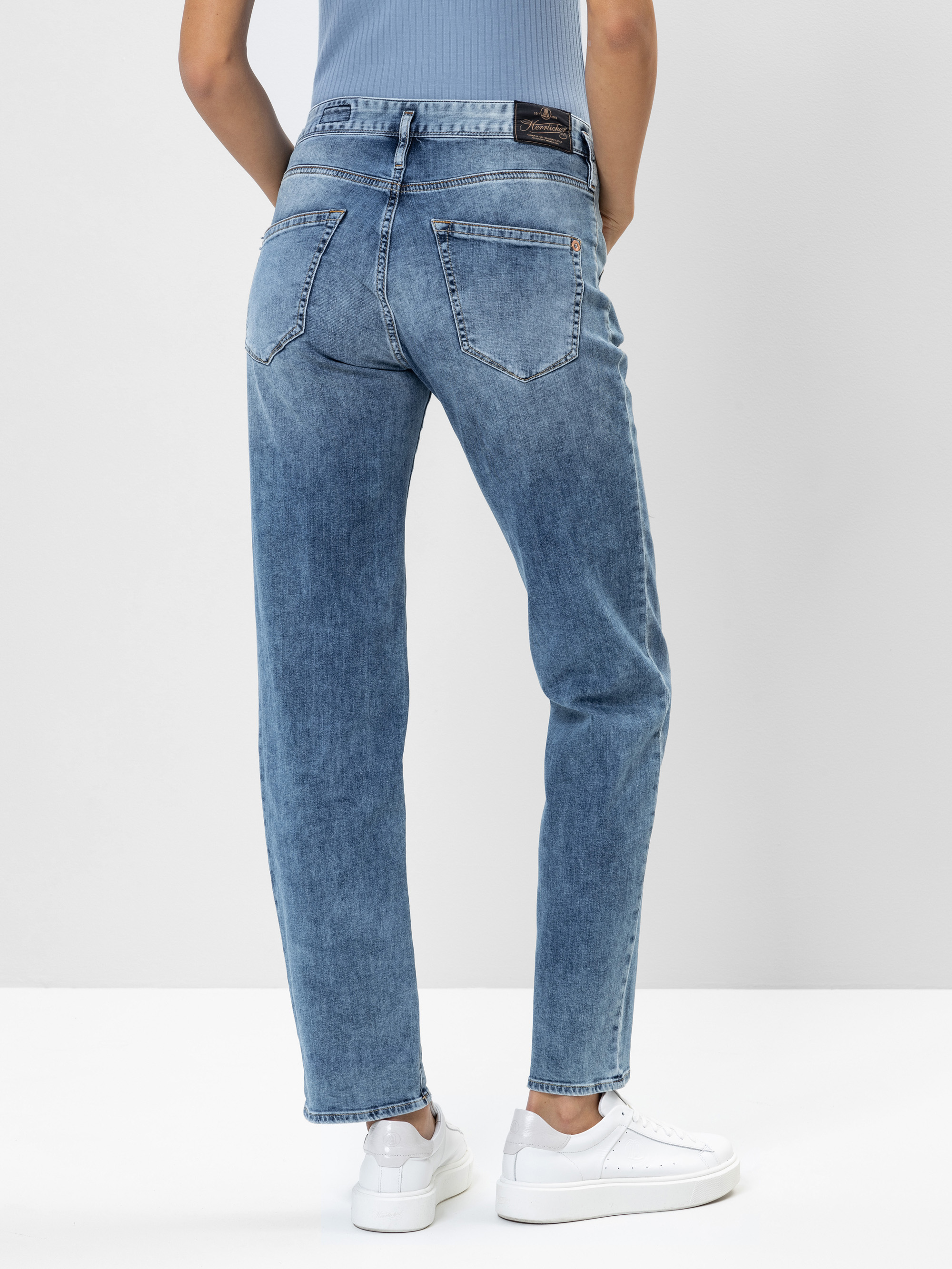 Mit dem Rücken zur Kamera stehende Person in hellblauen High-Waisted-Jeans und weißen Turnschuhen vor einem schlichten hellen Hintergrund.