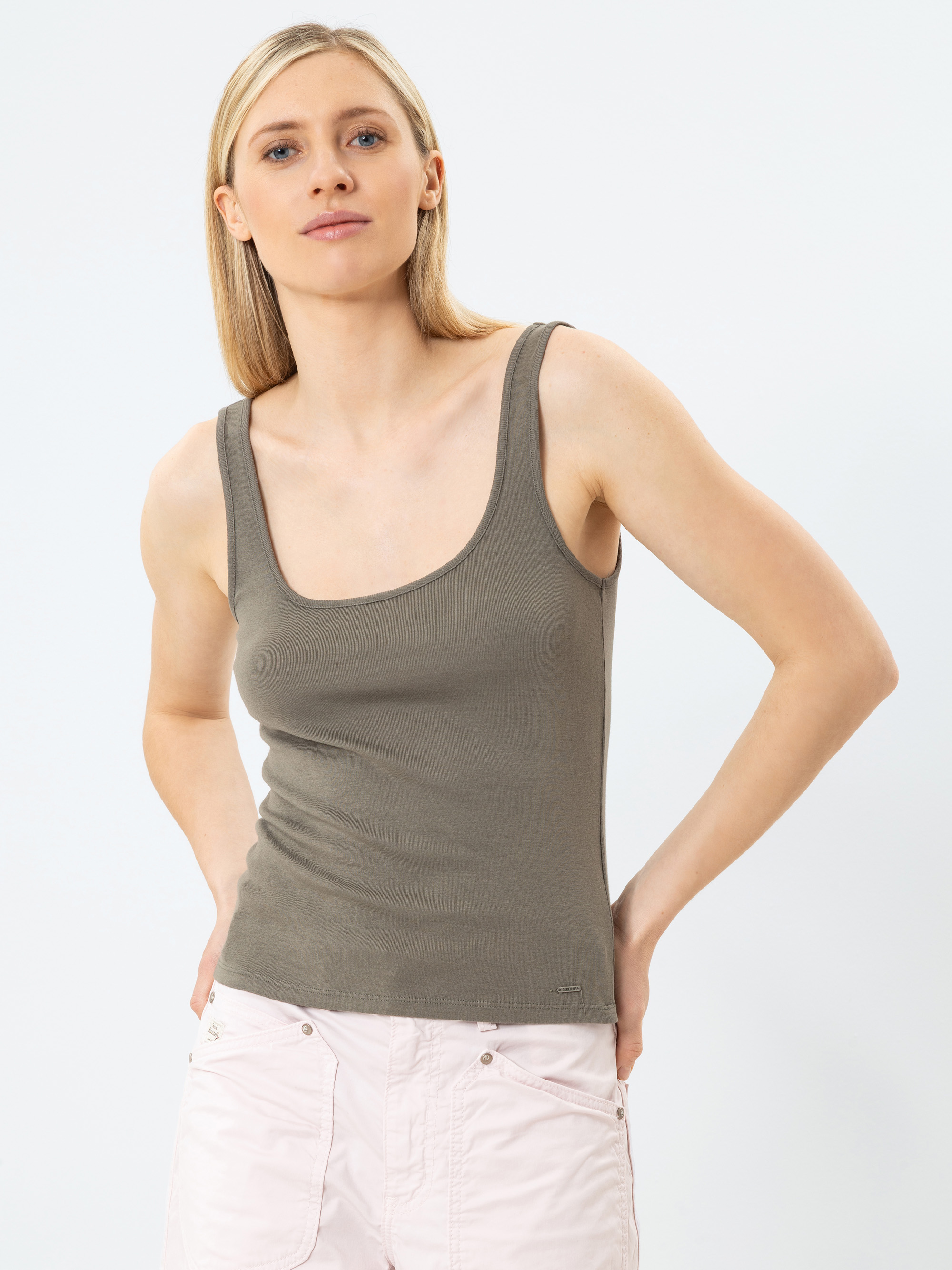 Eine Frau mit glattem blondem Haar trägt ein tailliertes, olivgrünes Tank-Top und eine hellrosa Hose. Sie steht vor einem schlichten weißen Hintergrund und blickt selbstbewusst in die Kamera, die Hände in die Hüften gestemmt.