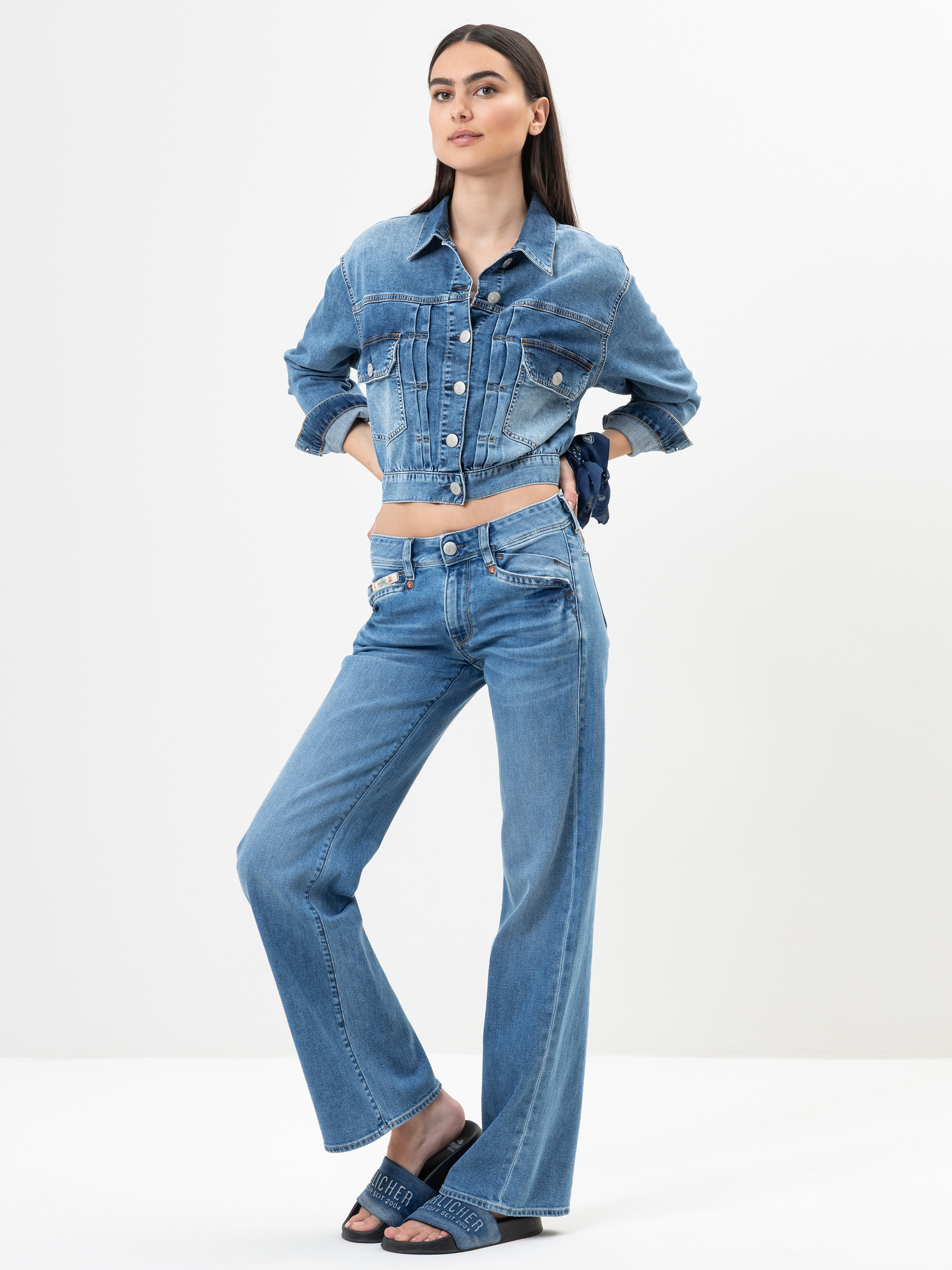 Eine Frau steht vor einem weißen Hintergrund und trägt eine Jeansjacke und hochgeschnittene Schlagjeans. Sie hat langes dunkles Haar, die Hände in den Taschen und trägt dunkle Slide-Sandalen.