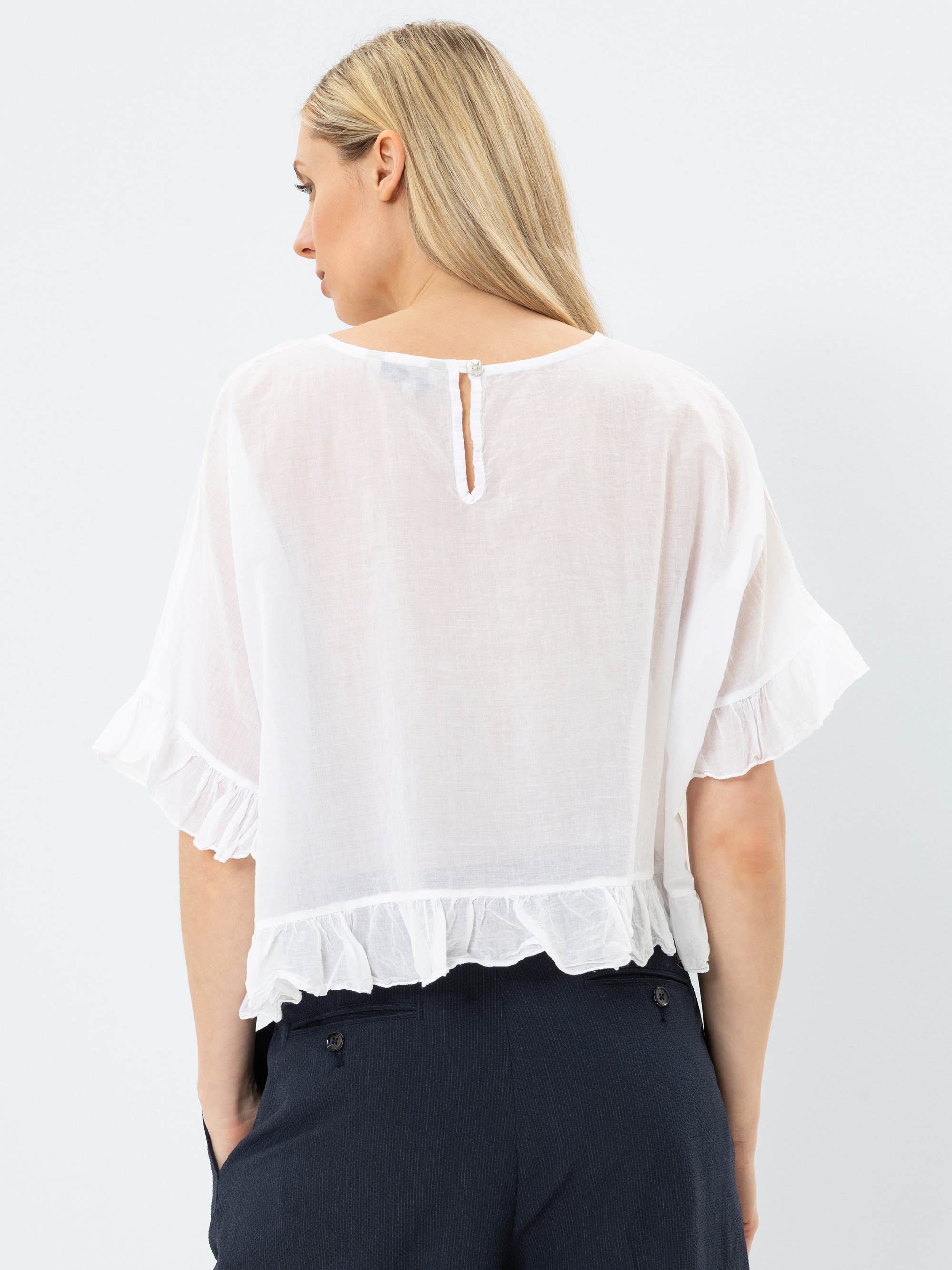 Eine Frau mit langen blonden Haaren trägt eine weiße kurzärmelige Bluse mit Rüschen und einem Schlüsselloch-Knopfverschluss auf der Rückseite, kombiniert mit einer dunklen Hose mit hoher Taille. Sie schaut von der Kamera weg.