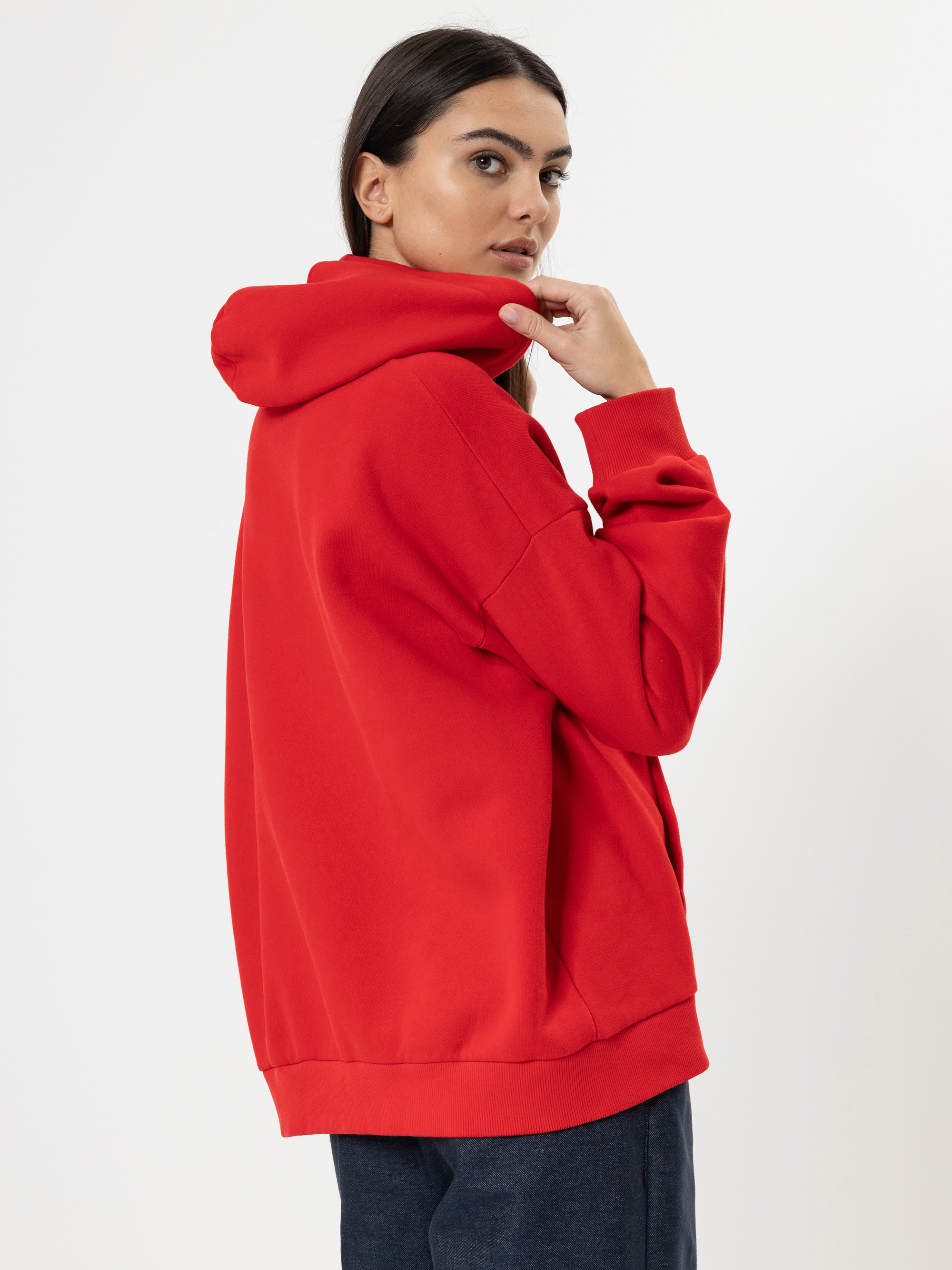 Eine Frau mit langen dunklen Haaren, die einen leuchtend roten Kapuzenpulli und eine dunkle Hose trägt, steht seitlich und blickt mit der Hand am Kinn vor einem schlichten weißen Hintergrund über ihre Schulter.