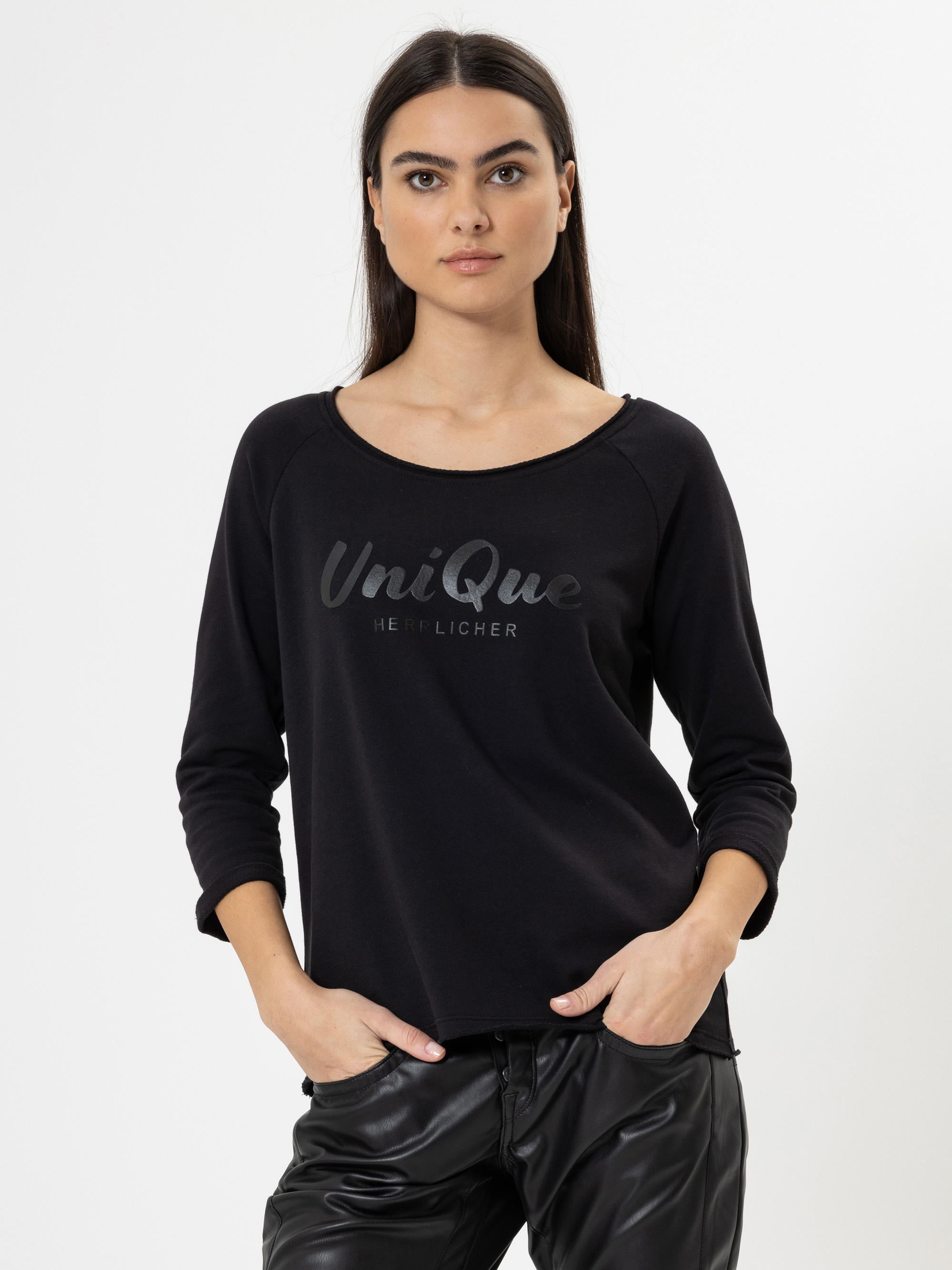 Eine Frau mit langen dunklen Haaren trägt ein schwarzes langärmeliges Hemd mit der Aufschrift Unique auf der Vorderseite und eine schwarze Lederhose. Sie steht mit den Händen in den Taschen vor einem schlichten weißen Hintergrund.