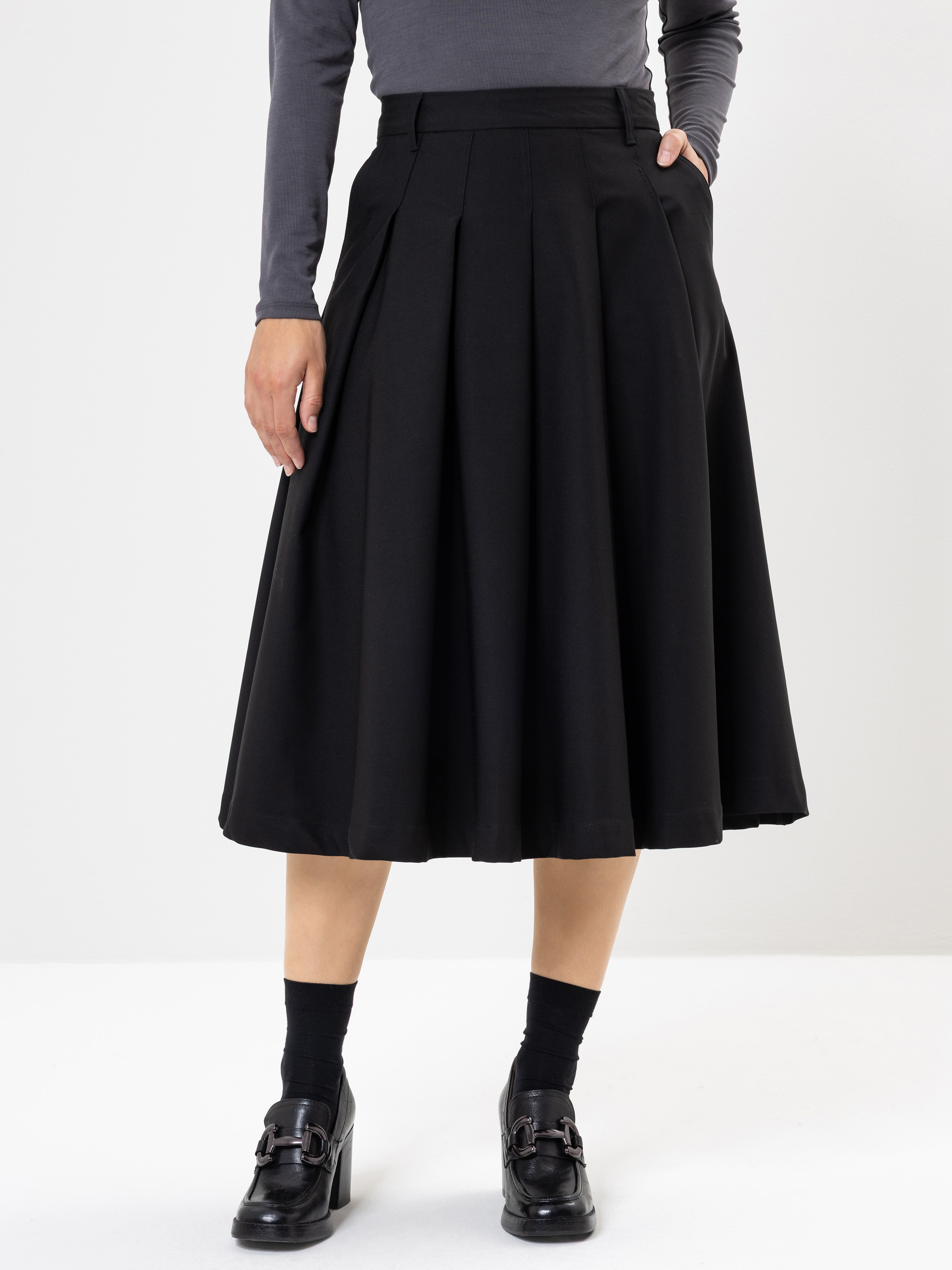 Eine Person, die ein langärmeliges, dunkelgraues Oberteil und einen hochgeschlossenen, knielangen schwarzen Faltenrock mit schwarzen Halbschuhen und schwarzen Socken trägt, steht vor einem schlichten weißen Hintergrund.